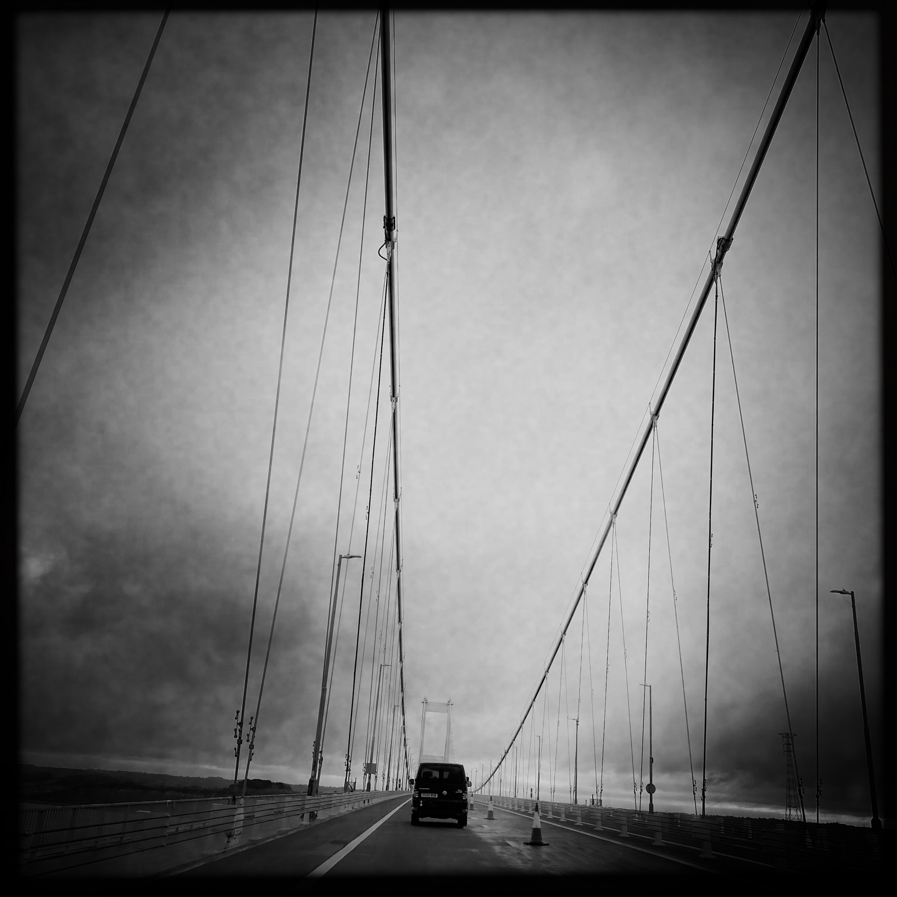 Black and white photo of a suspension bridge, with cables converging towards the top of the towers, and a single van driving along the road.