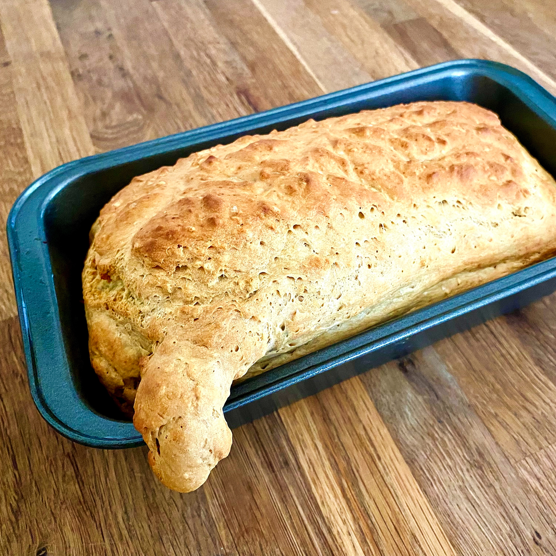A loaf of bread in a metal baking pan on a wooden surface. One corner of the loaf has risen over the corner and out of the tin.