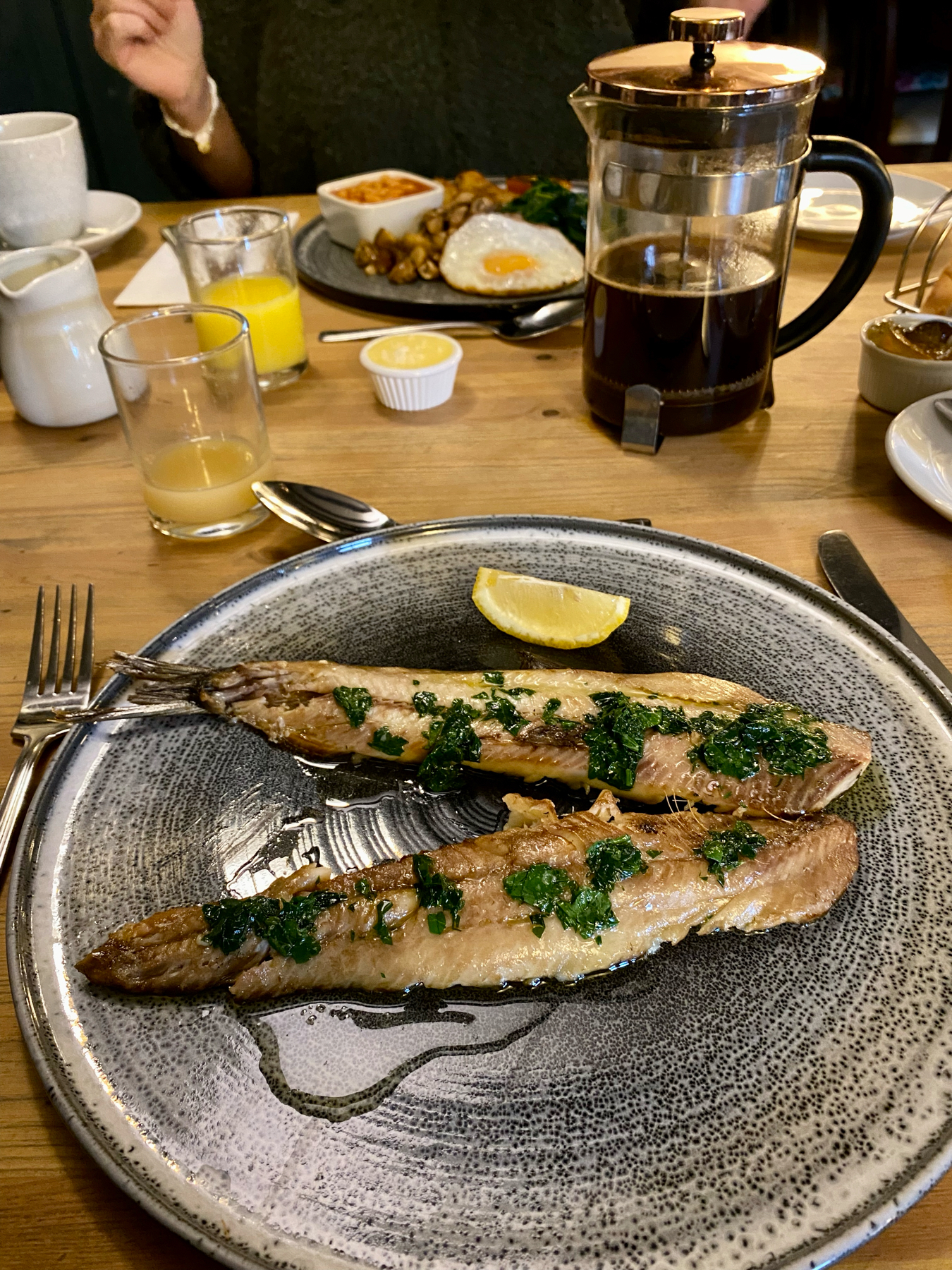 A breakfast table with two kipper fillets garnished with herbs on a plate, accompanied by a French press with coffee, orange juice, and another plate with fried eggs, spinach, and beans in the background.