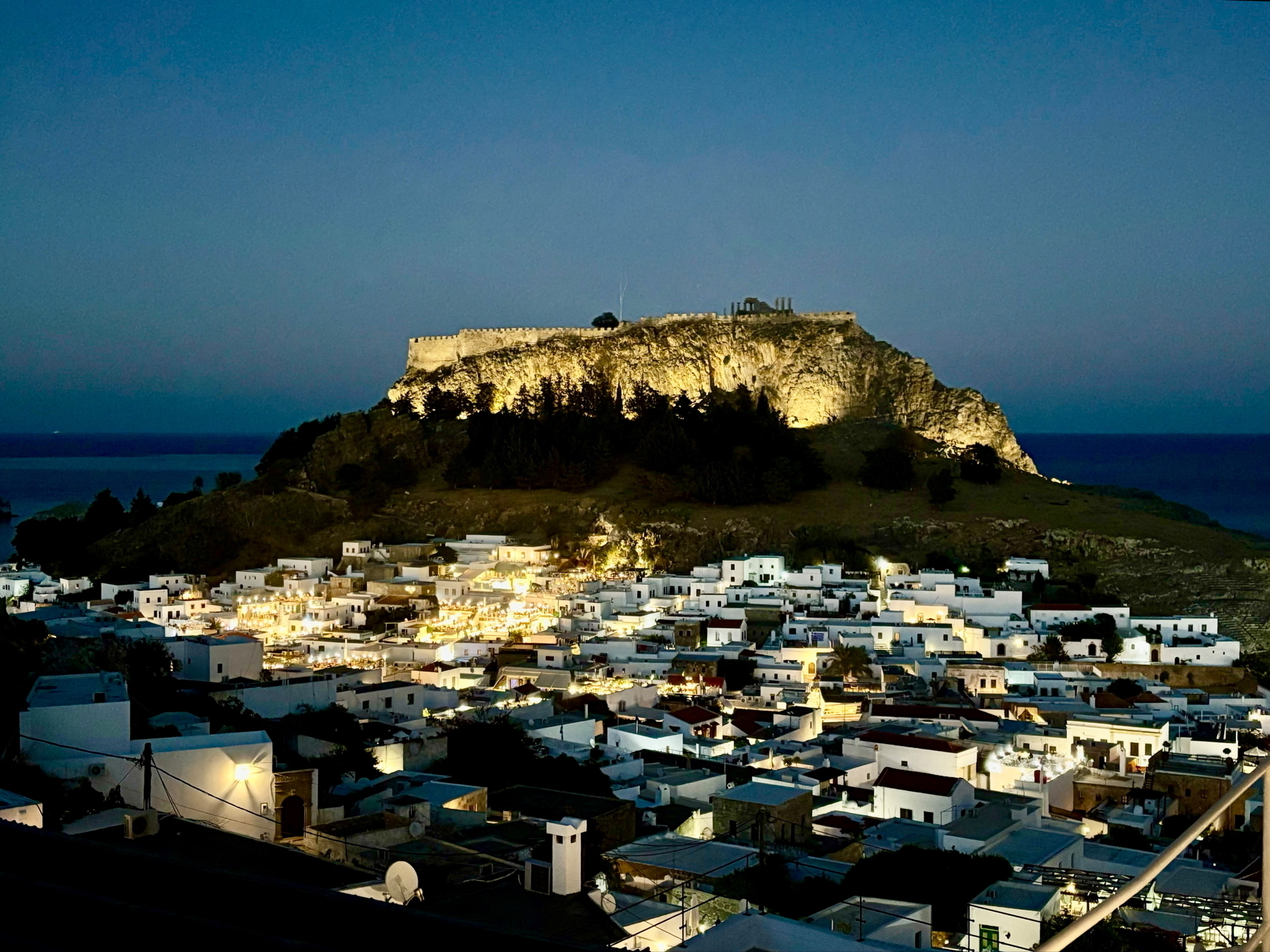 A nighttime view of a coastal village with white buildings, illuminated by warm lights. Above the village, a large, well-lit fortress sits on a hill with the sea visible in the background.