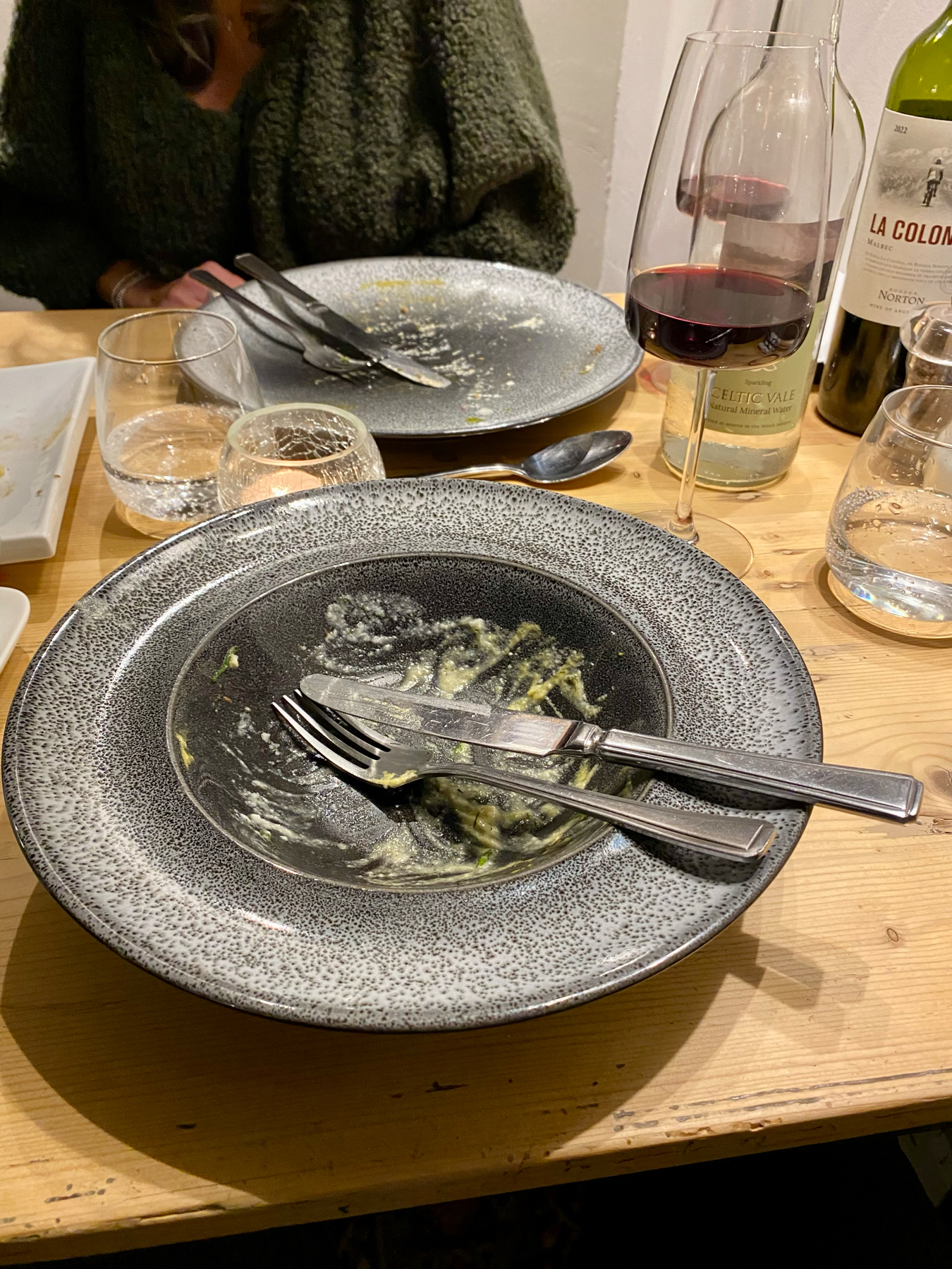 A nearly empty speckled dinner plate with remnants of food and a pair of crossed utensils on it, resting on a wooden table, surrounded by a half-empty wine glass, water glasses, and a wine bottle