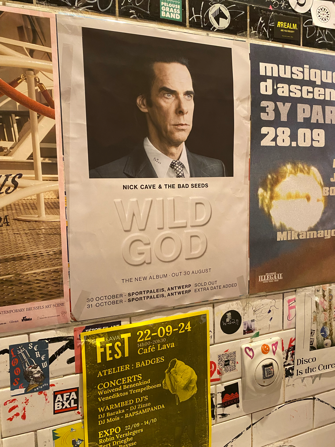 A wall cluttered with posters and stickers. The central poster promotes Nick Cave & The Bad Seeds and their new album Wild God set to release on August 30, with concert dates listed for October in Antwerp.