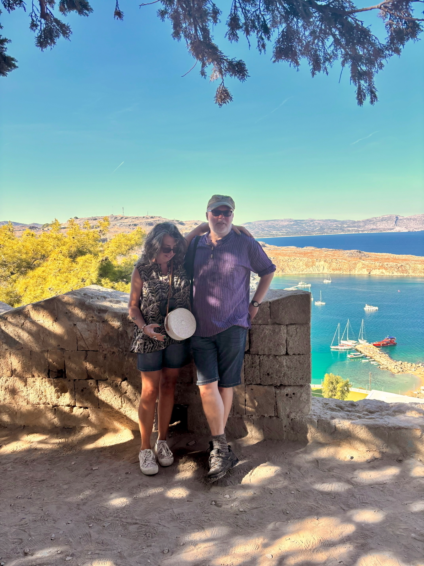 A couple is standing next to a stone wall overlooking a scenic coastal view. The woman is holding a round bag and wearing a patterned top and shorts, while the man is wearing a cap, sunglasses, and a striped shirt.