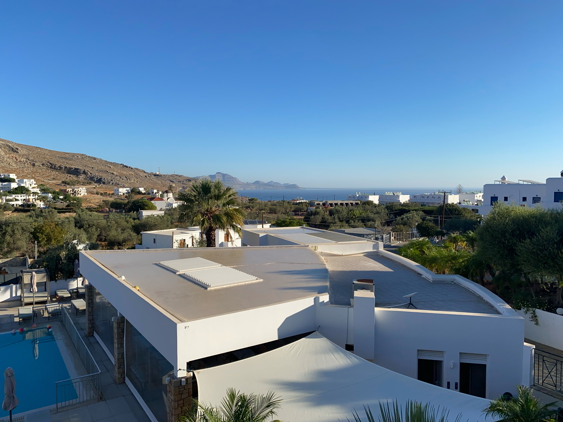 View of a Mediterranean coastal village with white buildings, a swimming pool, and lush greenery. A palm tree stands in the center, with distant mountains and the sea in the background under a clear blue sky.