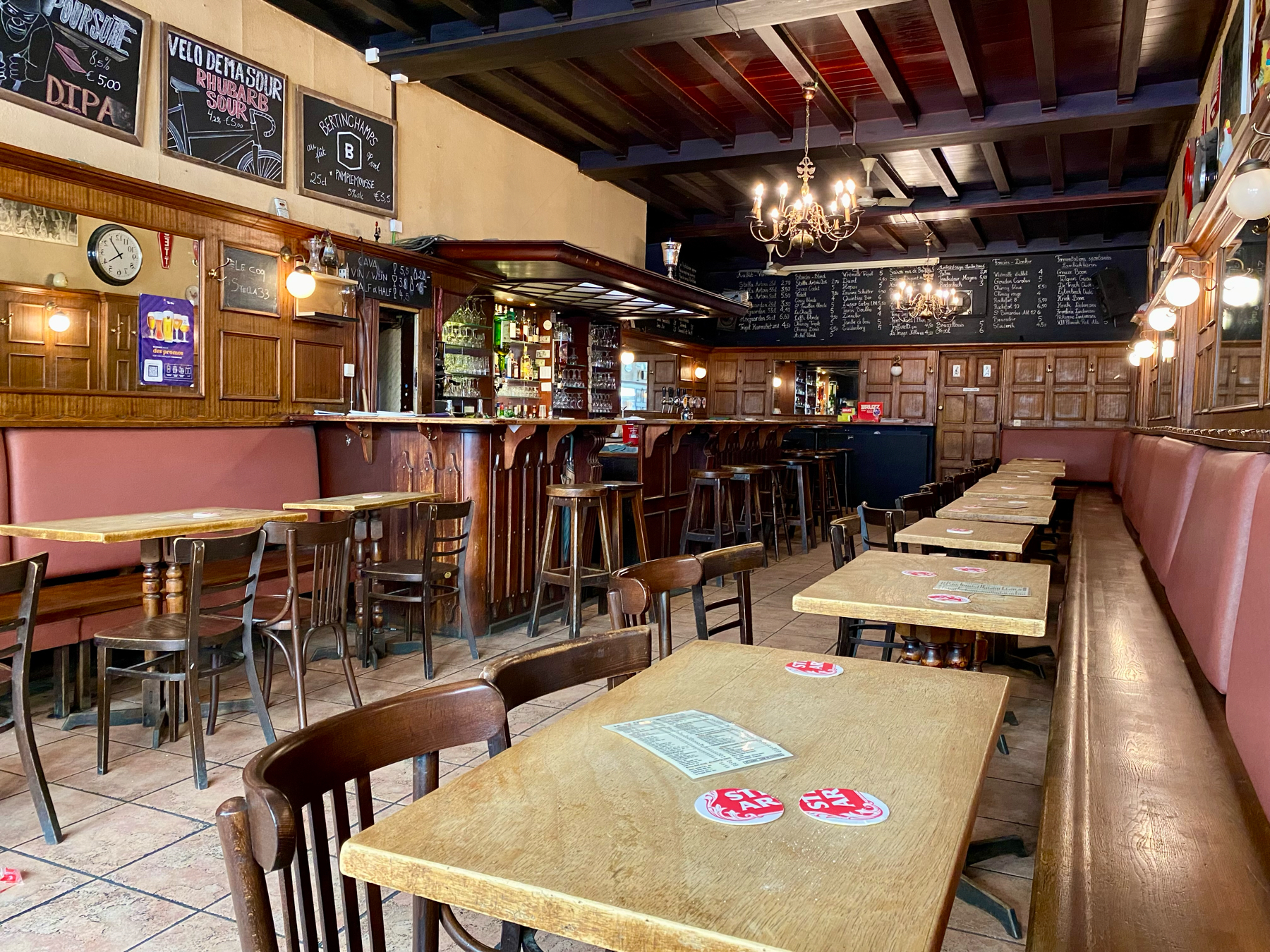 An empty, rustic pub interior with wooden tables and chairs, a long cushioned bench along the wall, and a wooden bar. The pub is adorned with chalkboards listing drinks, hanging light fixtures, and a dark wooden ceiling.