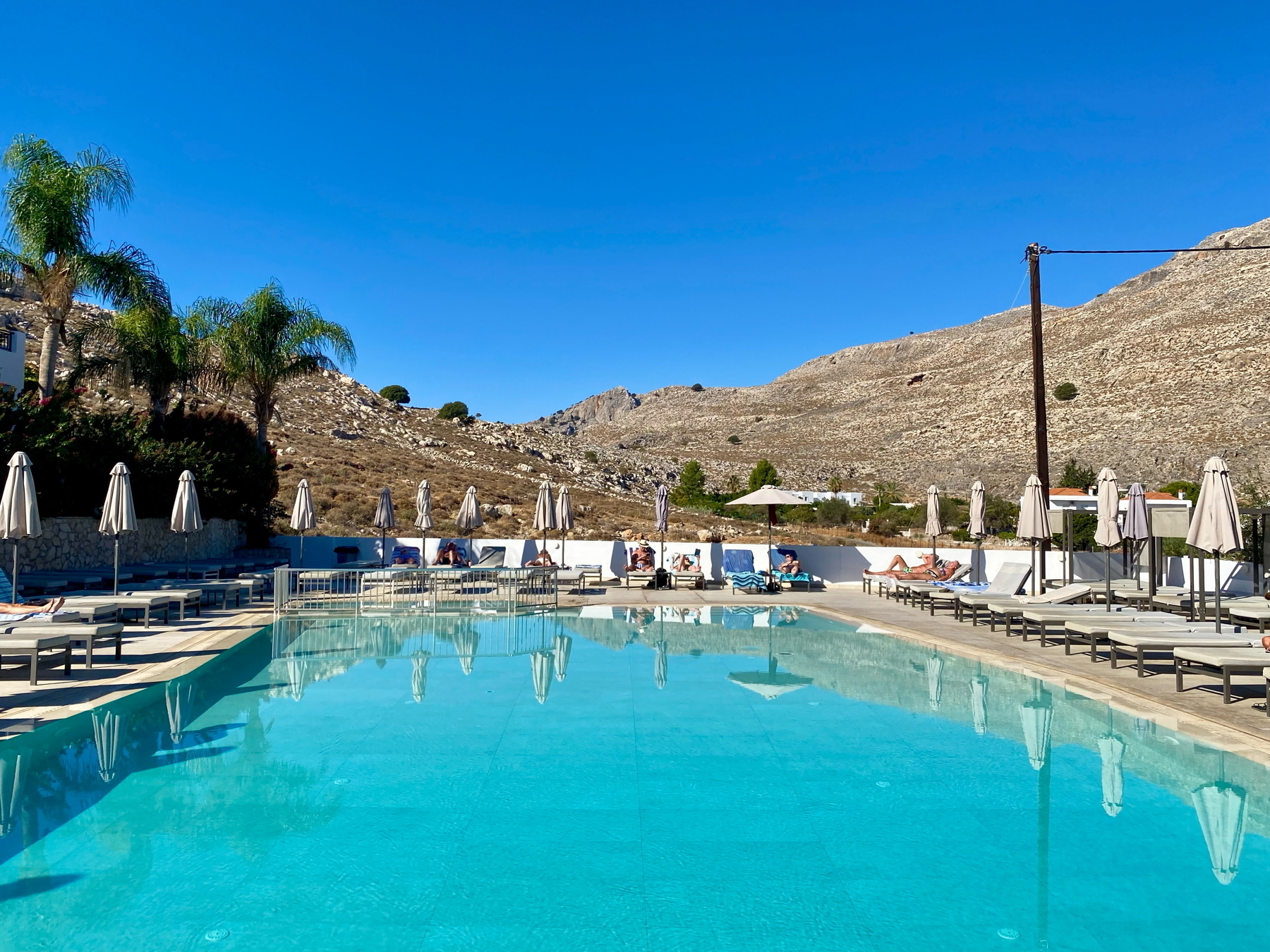 Outdoor swimming pool with clear blue water, surrounded by sunbeds and closed umbrellas. Palm trees and rocky hills are in the background under a clear blue sky.