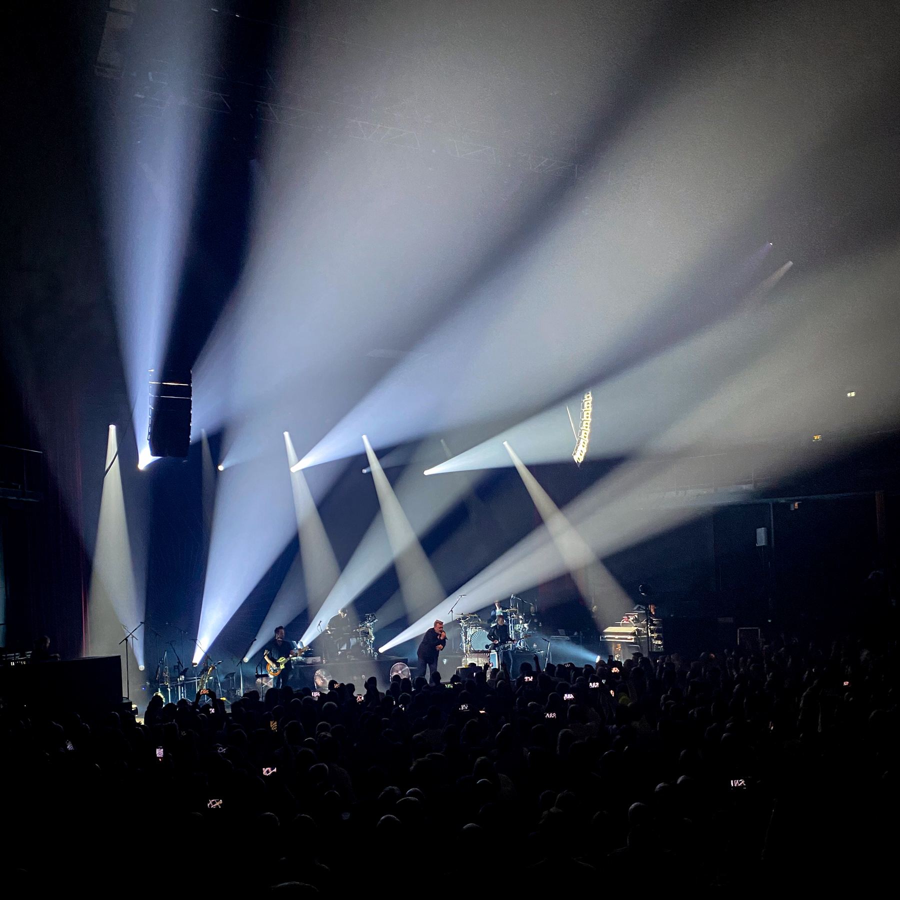 Concert scene with dramatic lighting and beams of light shining onto the stage. A band is performing, with musicians playing instruments and a vocalist singing. A large audience watches, many holding up their phones to record the event.