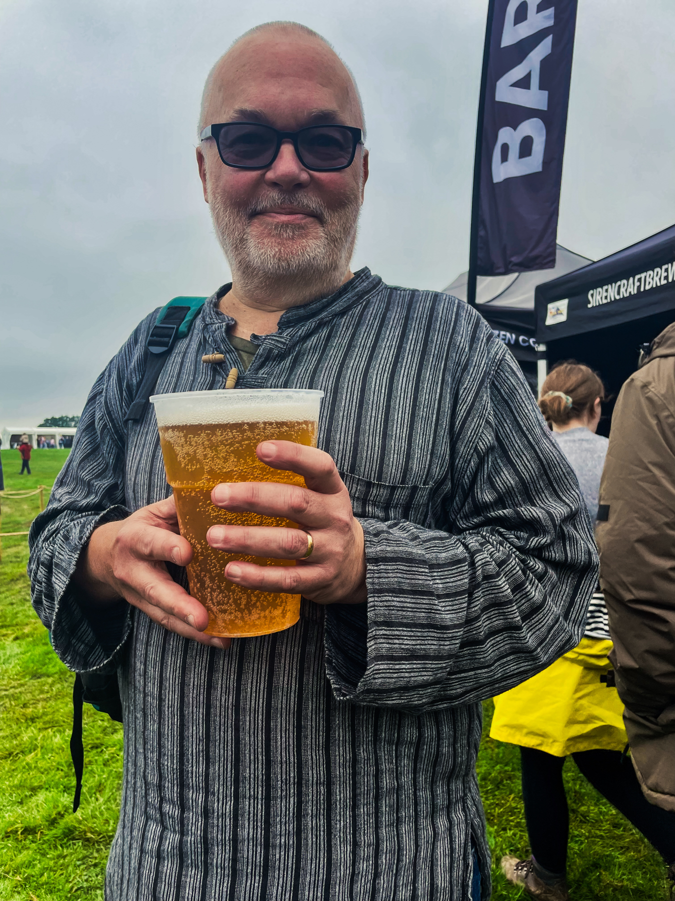 A man with a beard and glasses is holding a large cup of beer at an outdoor event. He is wearing a striped shirt and has a backpack on one shoulder. There are tents and other people in the background, with a banner that says BAR.