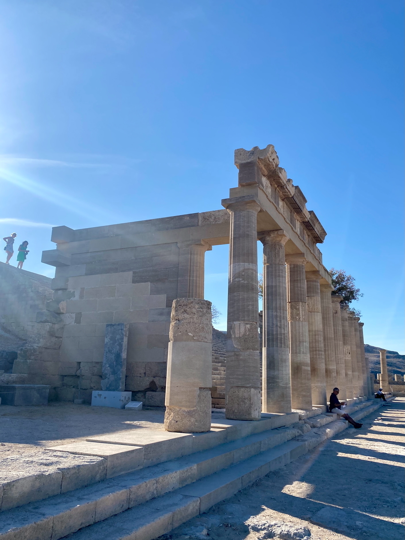 Ancient Greek ruins with tall columns and stone steps under a clear blue sky. Two people are sitting on the steps, and a few figures are visible on a higher level in the background.