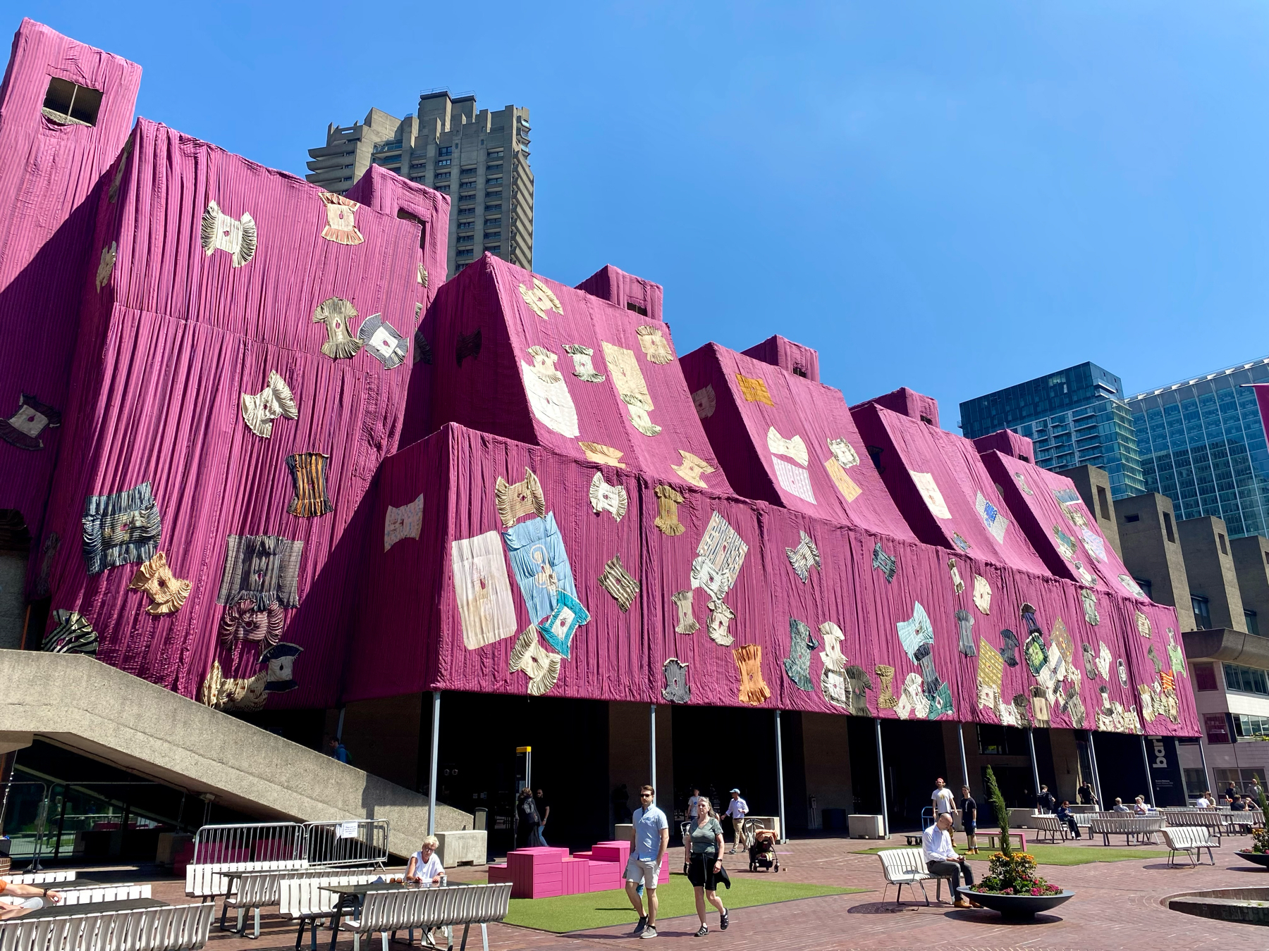 A large building is wrapped in magenta fabric adorned with various textile art pieces. The surrounding area features people walking, sitting on benches, and a few outdoor tables in a plaza on a sunny day. Modern skyscrapers are visible in the background.