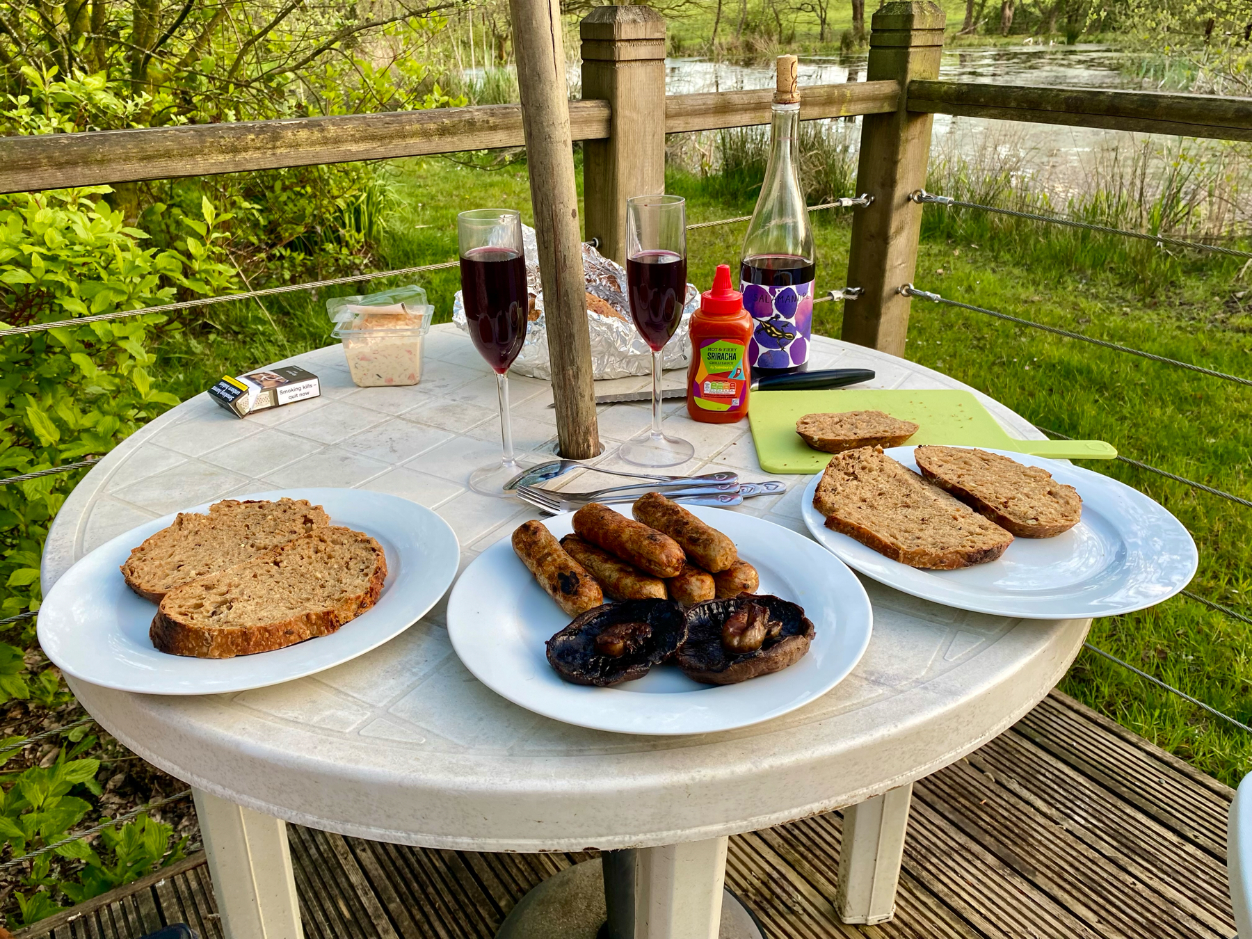 Outdoor meal setup with two glasses of red wine, a bottle, grilled sausages, slices of bread on plates, coleslaw, Sriracha sauce, and condiments on a white round table with a natural backdrop.