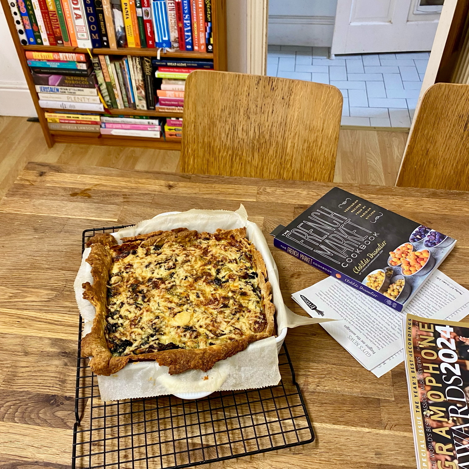 A baked tart on a cooling rack sits on a wooden table. Next to it is an open cookbook titled The French Market Cookbook. In the background is a bookshelf filled with cookbooks.