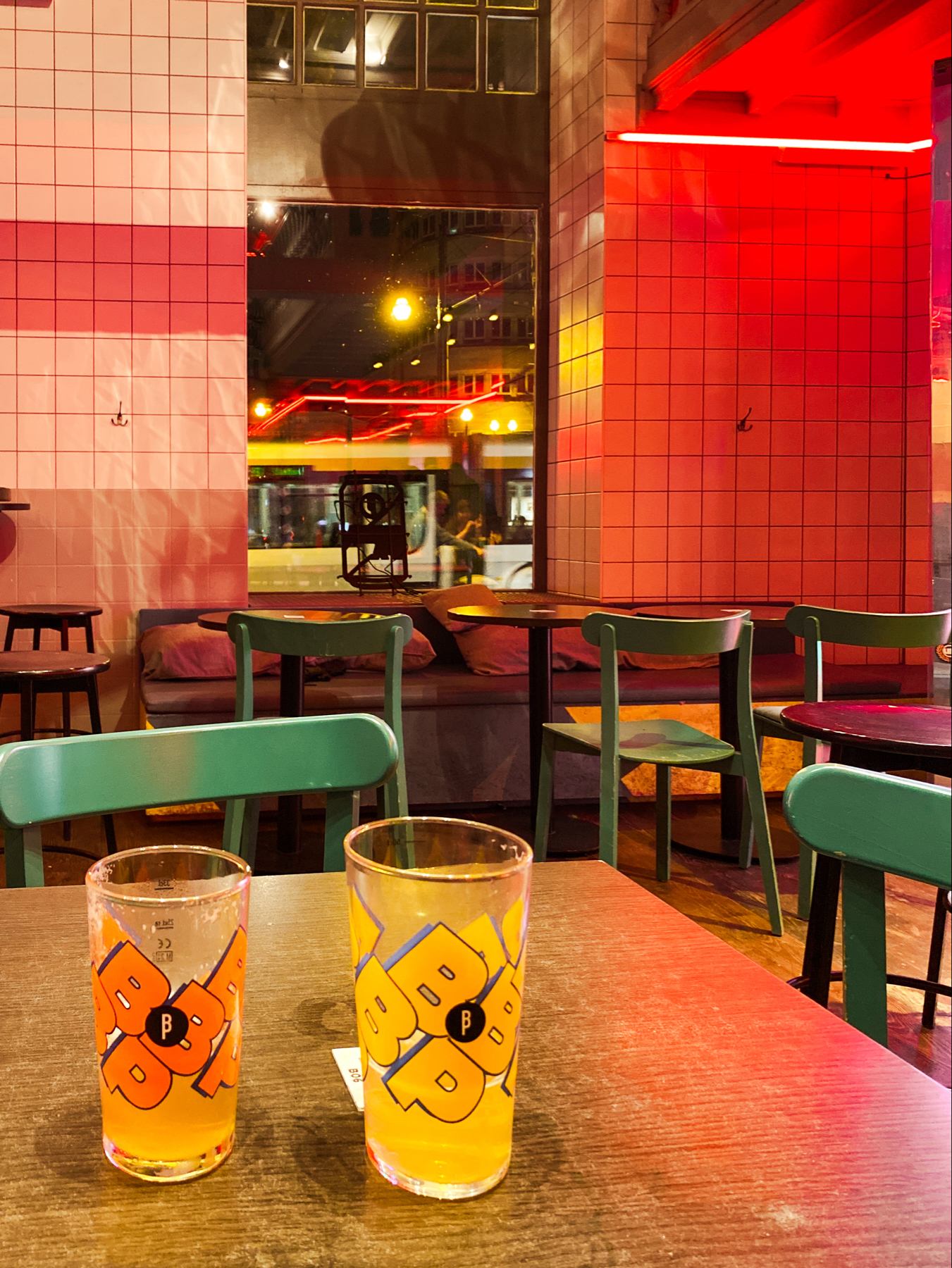 An indoor café or bar scene with a wooden table and two glasses of beer in the foreground. The glasses have colorful designs on them. The seating includes green chairs, and the walls are decorated with white and red tiles. There is red neon lighting illuminating the scene.