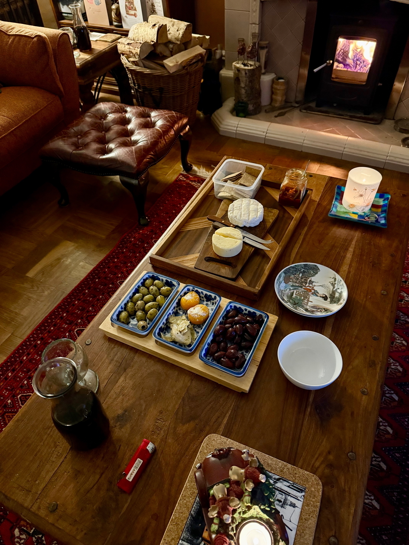 A cozy living room features a wooden coffee table with an assortment of cheeses, olives, and snacks, set against the backdrop of a lit fireplace.