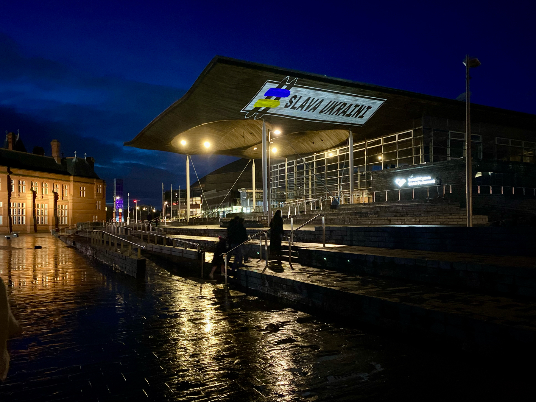 Night scene of the exterior of the Senedd, also known as the Welsh Parliament building, in Cardiff Bay with the building illuminated and a wet promenade reflecting lights. People are visible walking by.