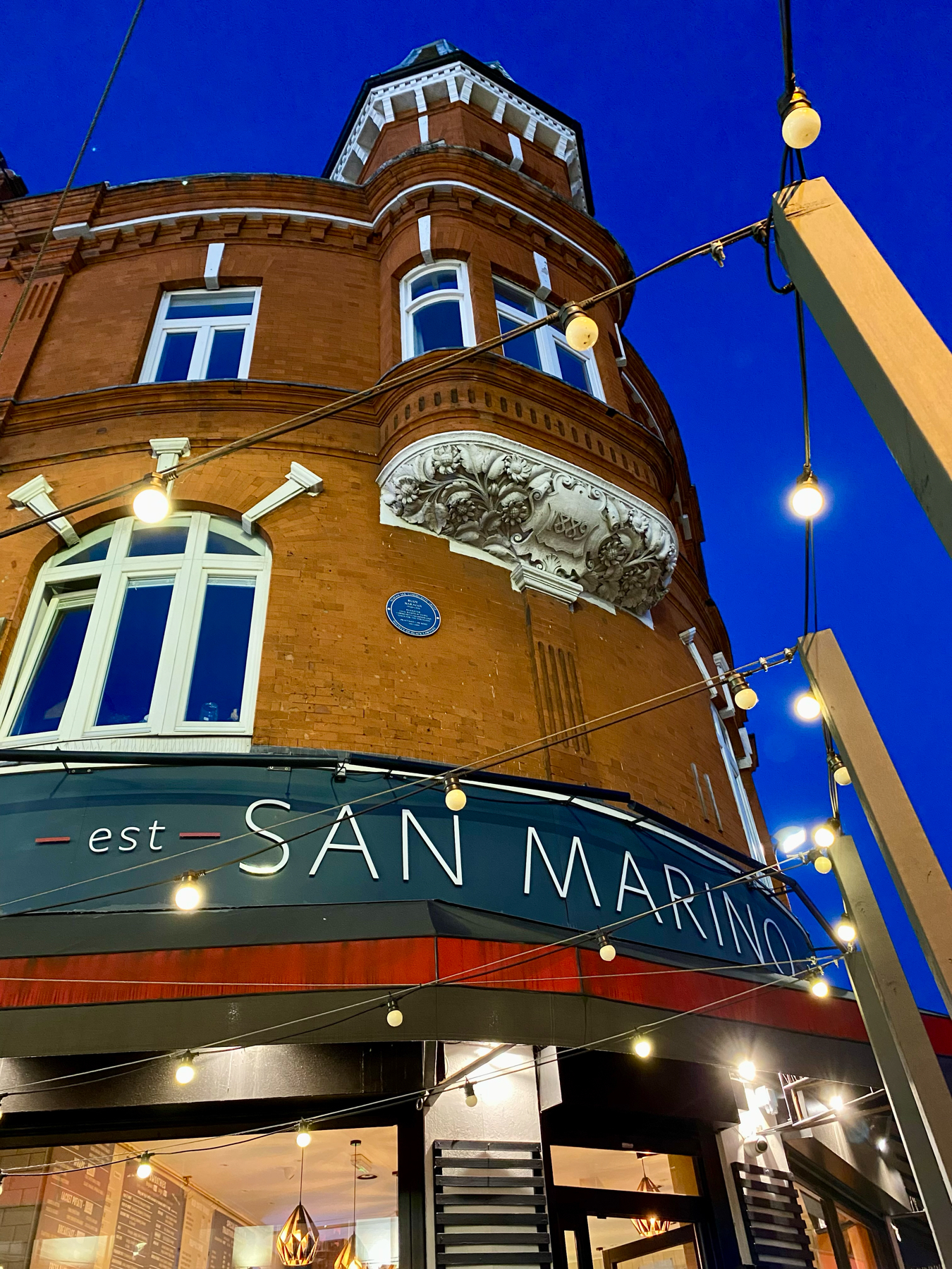 Image of a corner building with ornate architectural details, featuring a turret and large windows. The sign on the building reads San Marino. The scene is illuminated with string lights hanging across the storefront, against a backdrop of a deep blue evening sky.