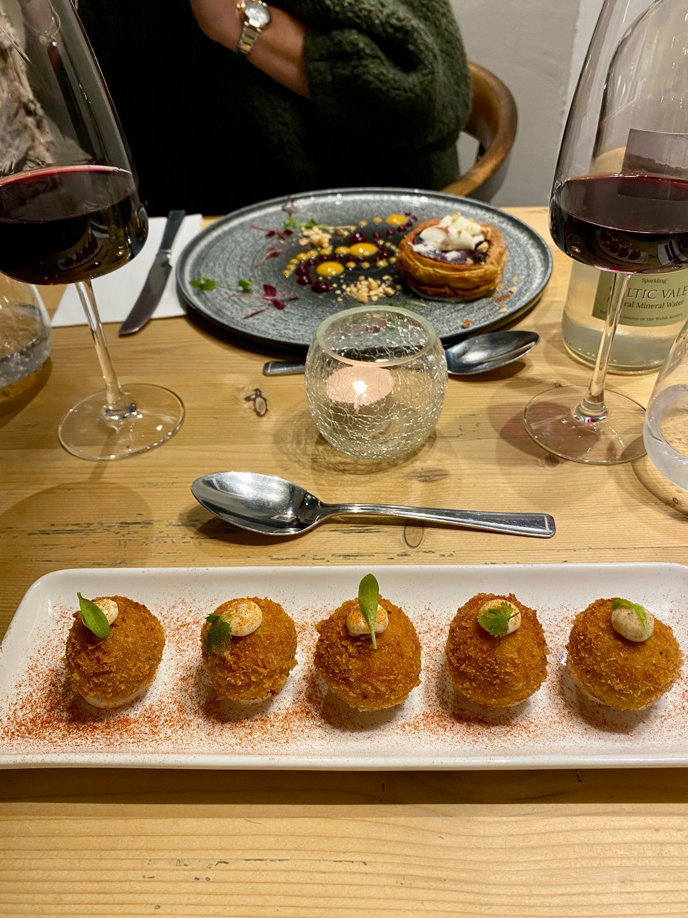 A table with gourmet dishes and wine glasses; in the foreground, a plate with breaded appetizers garnished with green leaves, sprinkled with paprika; in the background, two glasses of red wine, another dish with colorful food