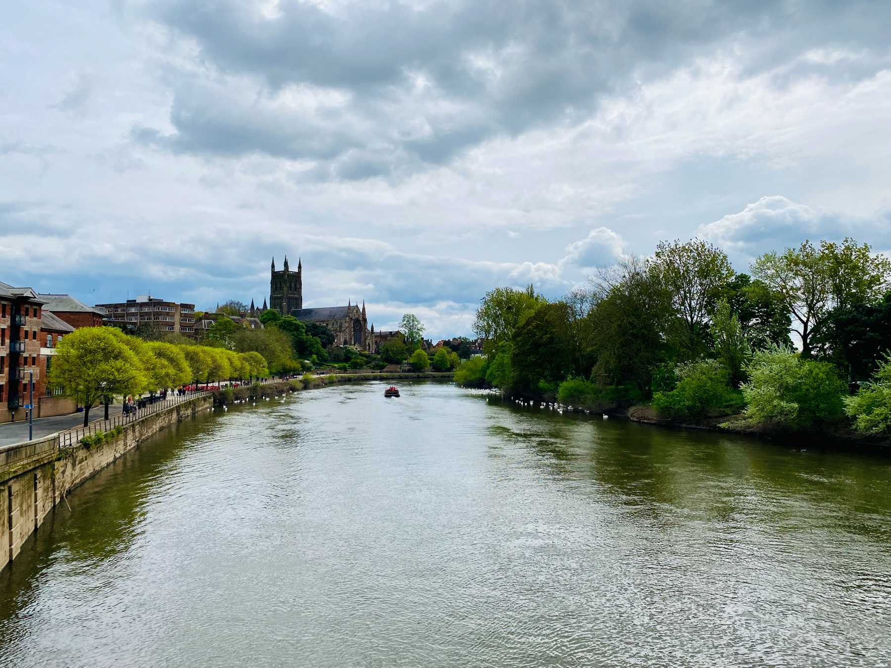 A river flowing through a city with green trees lining the banks, a cathedral in the distance, and a cloudy sky above.