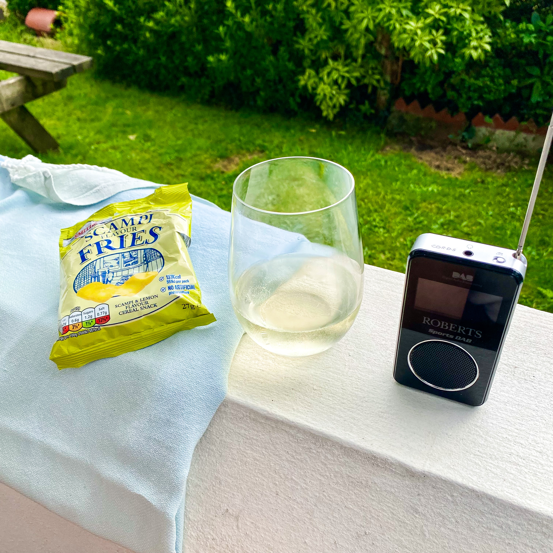 An outdoor scene featuring a white wall with a packet of Scampi Flavour Fries, a glass of white wine, and a Roberts sports DAB radio.