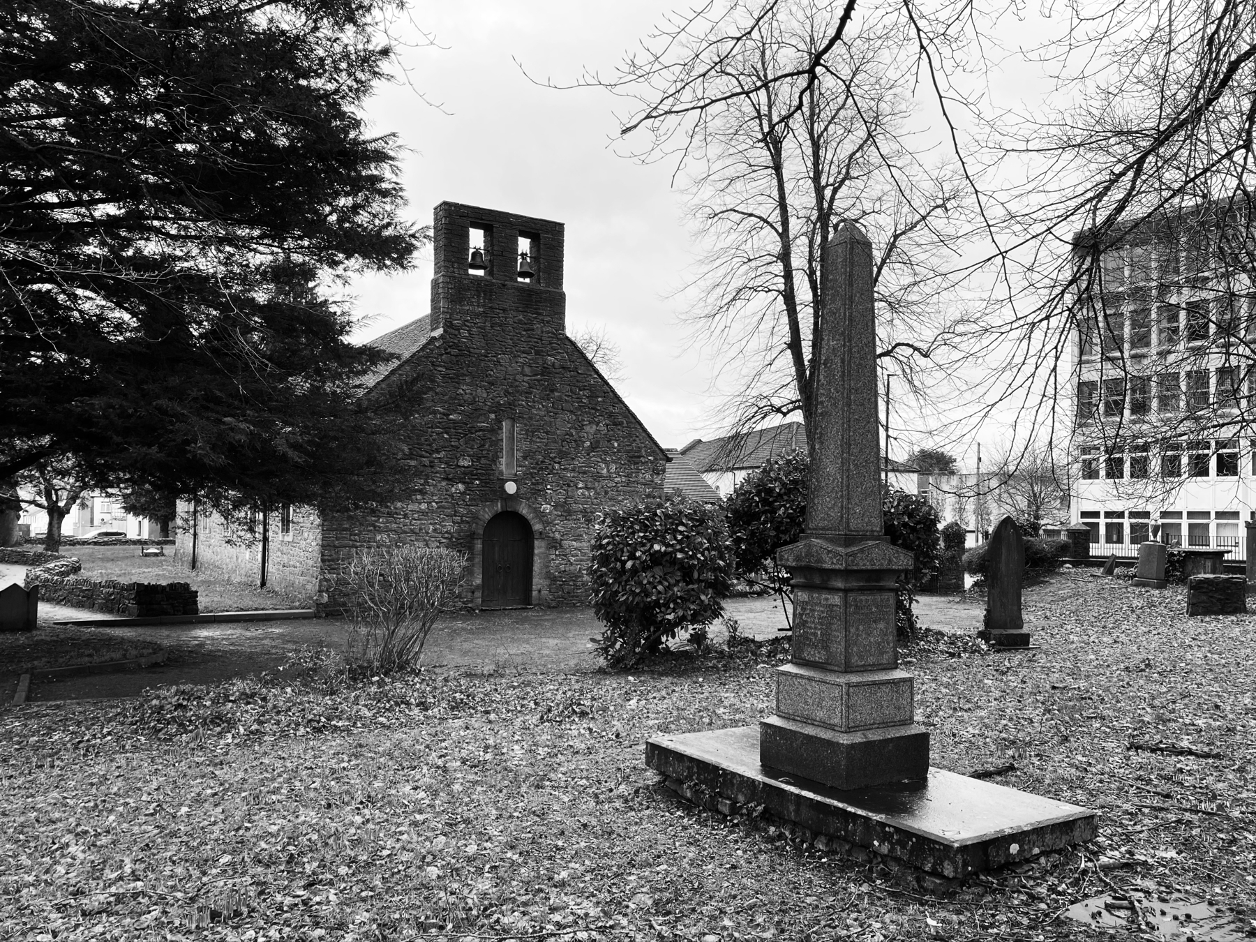 Black and white photo of an old stone chapel with a bell tower next to a cemetery with gravestones and a prominent memorial obelisk. Leafless trees and a modern building are in the background.