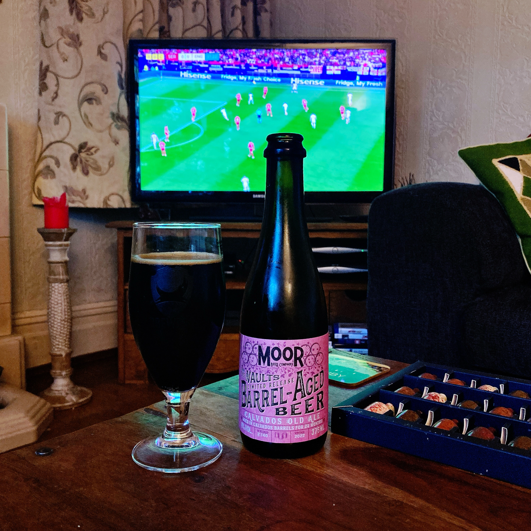 A glass and a bottle of Moor Vaults limited release barrel-aged beer are on a wooden coffee table. A tray of assorted chocolates is also on the table. In the background, a television screen shows a soccer match being played.