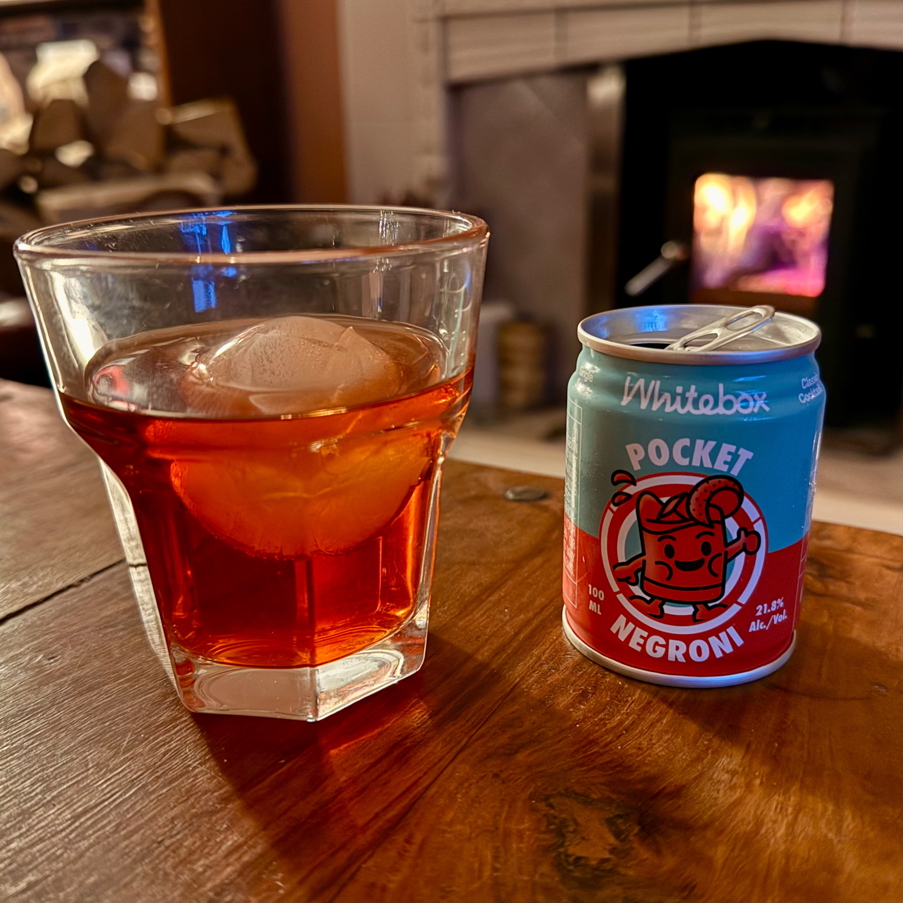 A glass with an ice cube contains a red cocktail next to a small can labeled Pocket Negroni on a wooden surface in a cozy setting.