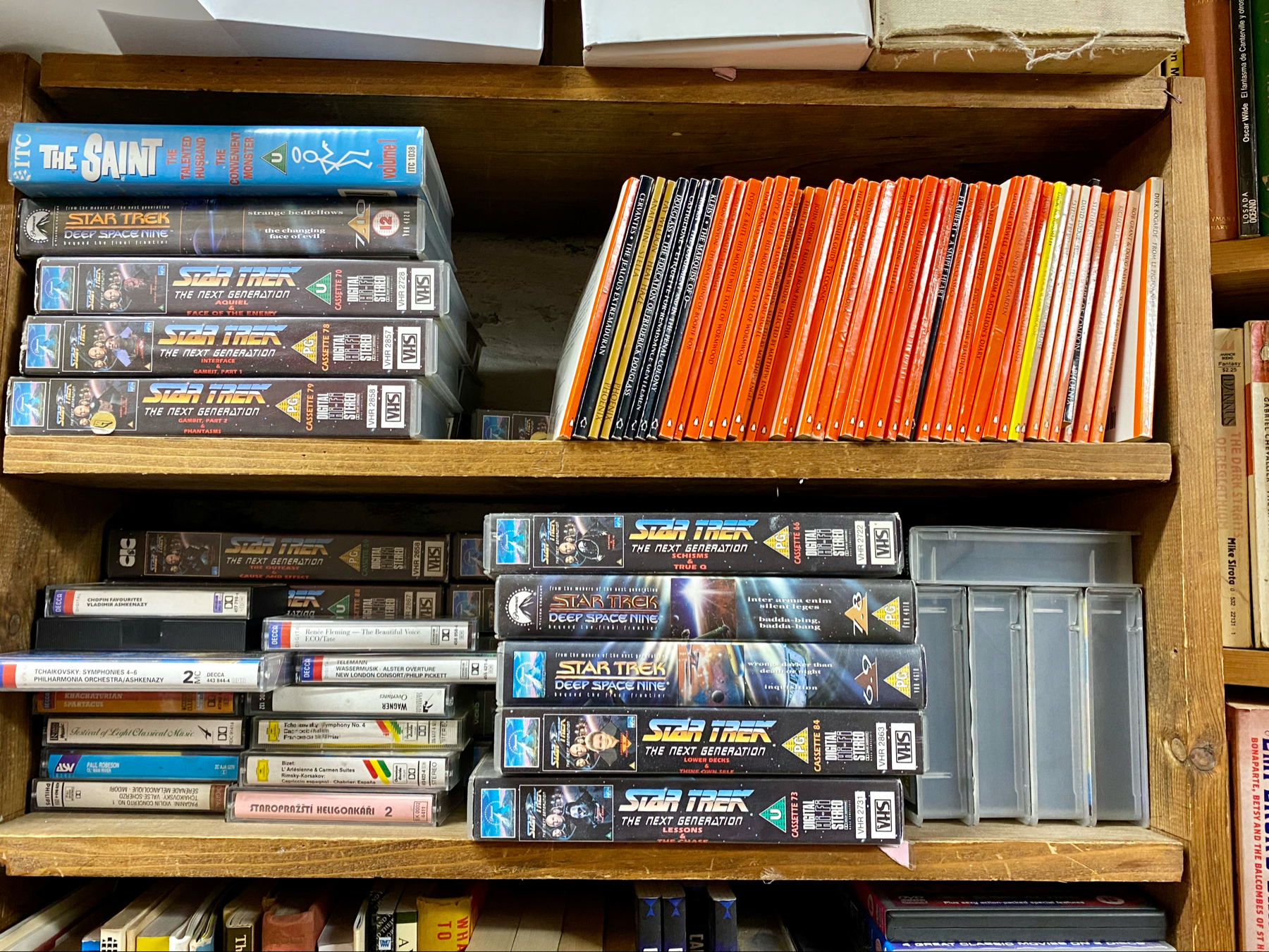 A wooden bookshelf filled with various VHS tapes, mostly from the Star Trek series, and a row of orange-spined books. The top shelf includes a box set for The Saint TV series.