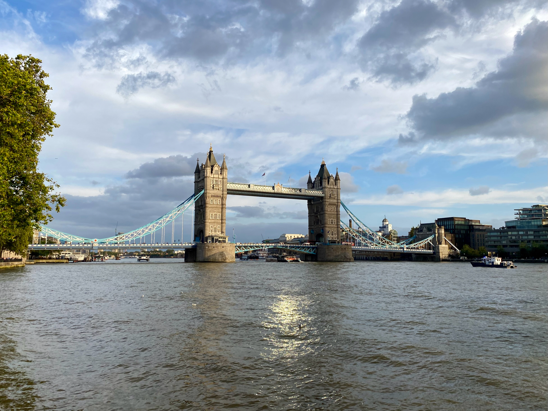 Tower Bridge in London spanning the River Thames on a cloudy day, with boats on the river and trees on the left bank.