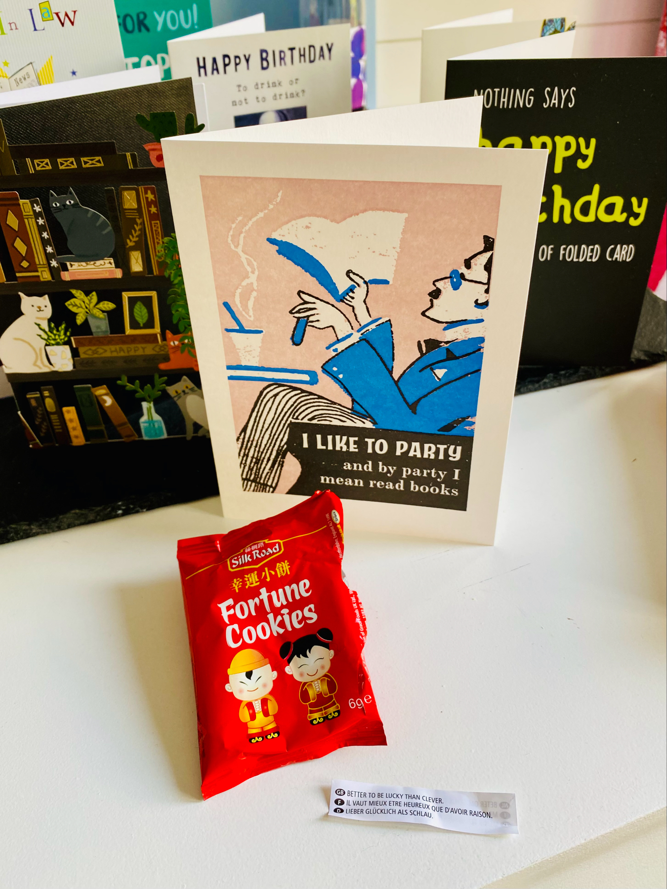 A collection of birthday greeting cards displayed on a surface, one featuring an illustration of a person reading with the phrase I like to party and by party I mean read books. In front of the cards, there's a fortune cookie wrapper