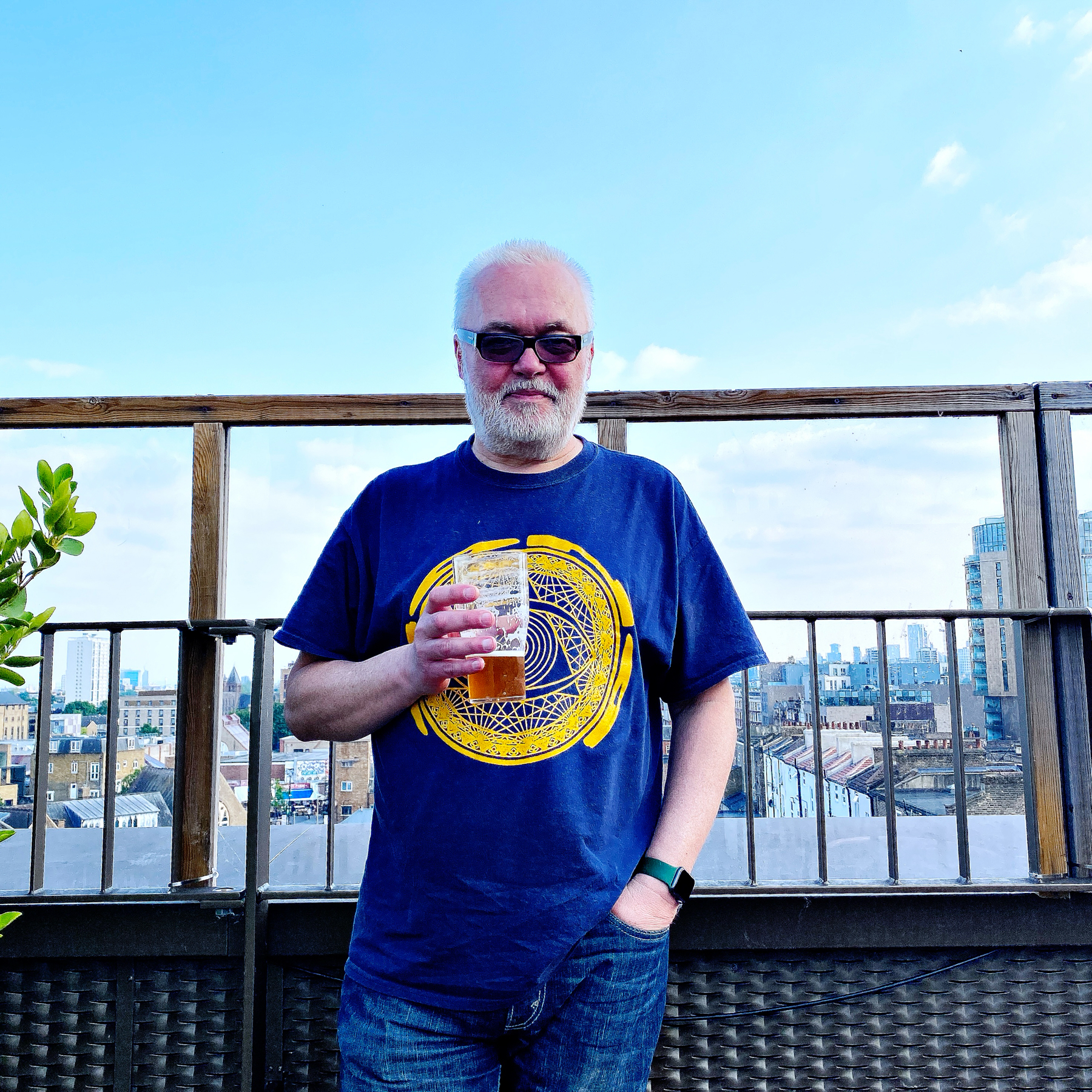 A man with white hair and beard is standing on a rooftop holding a pint of beer. He is wearing sunglasses and a blue T-shirt with a yellow circular design. The background includes a cityscape with buildings and a blue sky.