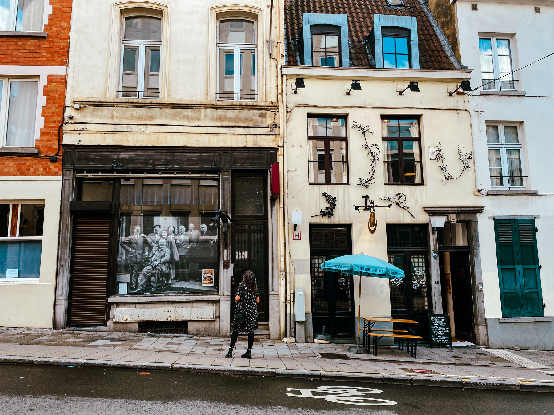 A street scene showing a row of buildings with large windows. The building on the left has a storefront with a large black-and-white photograph of people displayed in the window. The building on the right houses a café with decorative metalwork and blue accents.