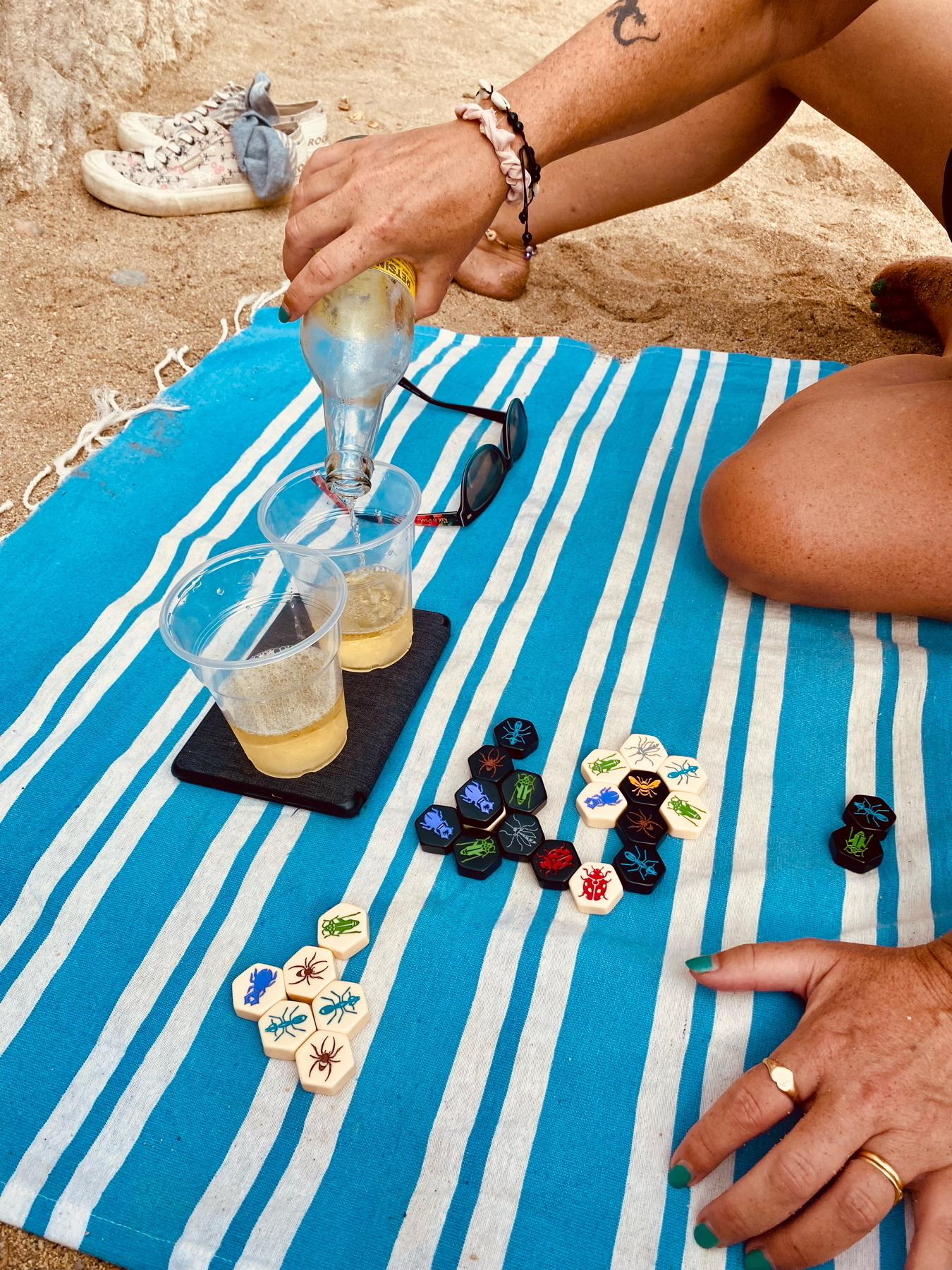 Two people playing a board game with hexagonal tiles featuring insect designs on a blue and white striped beach towel. One person is pouring white wine into plastic cups, and there are sunglasses and sneakers nearby.