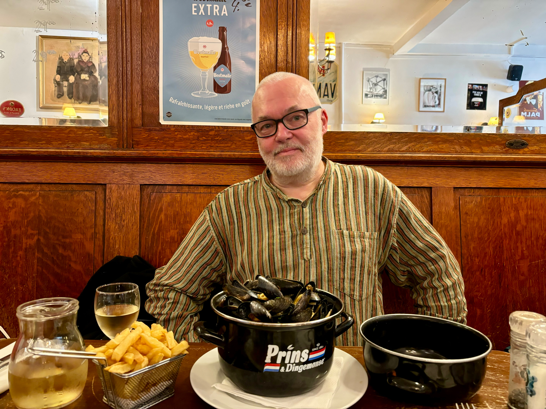 A man with glasses and a striped shirt is seated at a wooden table in a cozy restaurant. In front of him is a meal consisting of a pot of mussels, a basket of fries, and a glass of white wine. The background features wooden panels and mirrors.