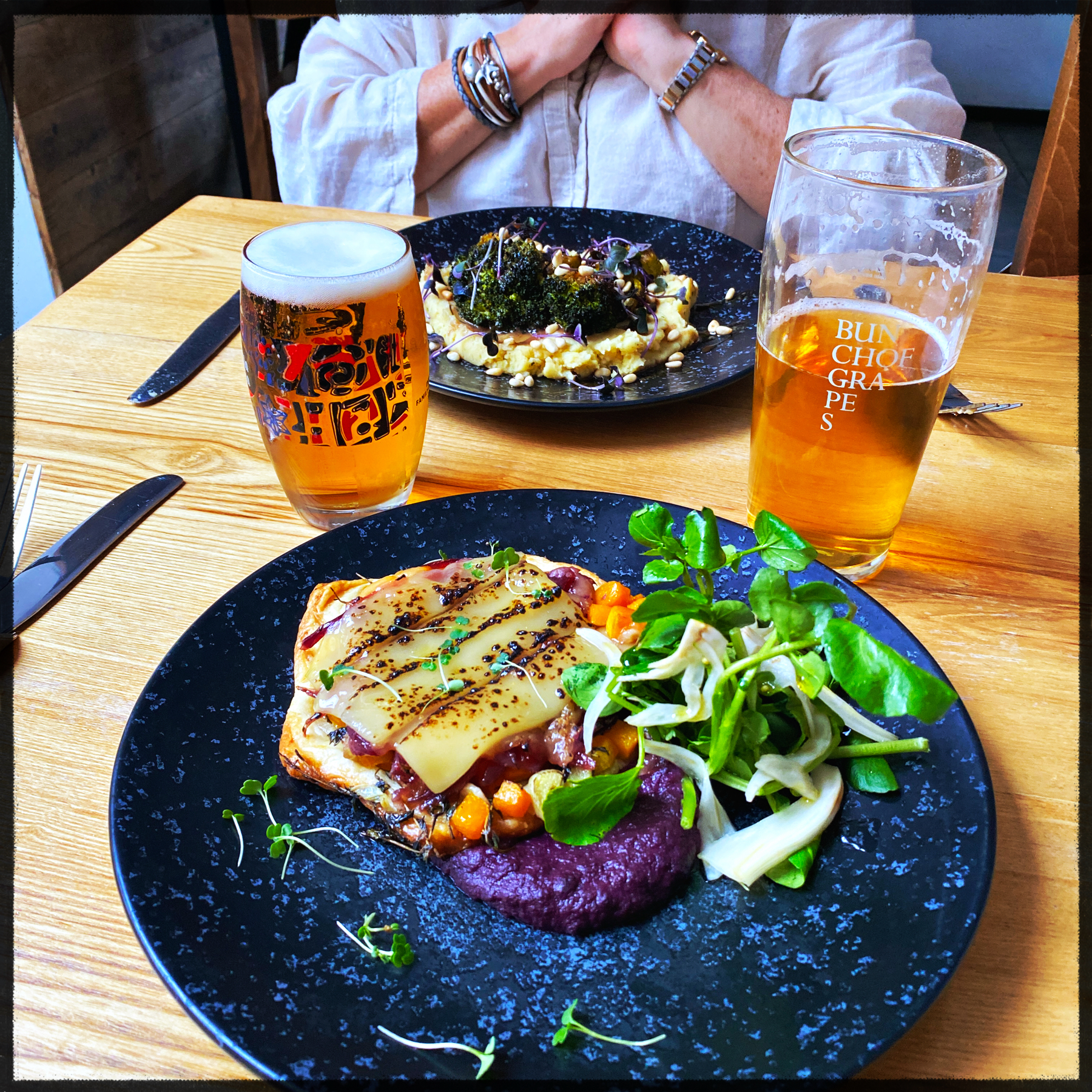 A wooden table setting at a restaurant with two plates of gourmet food and two glasses of beer. The foreground plate features a dish with cheese, a vegetable tart, and a side salad; the background plate has a different dish topped with grilled broccoli.