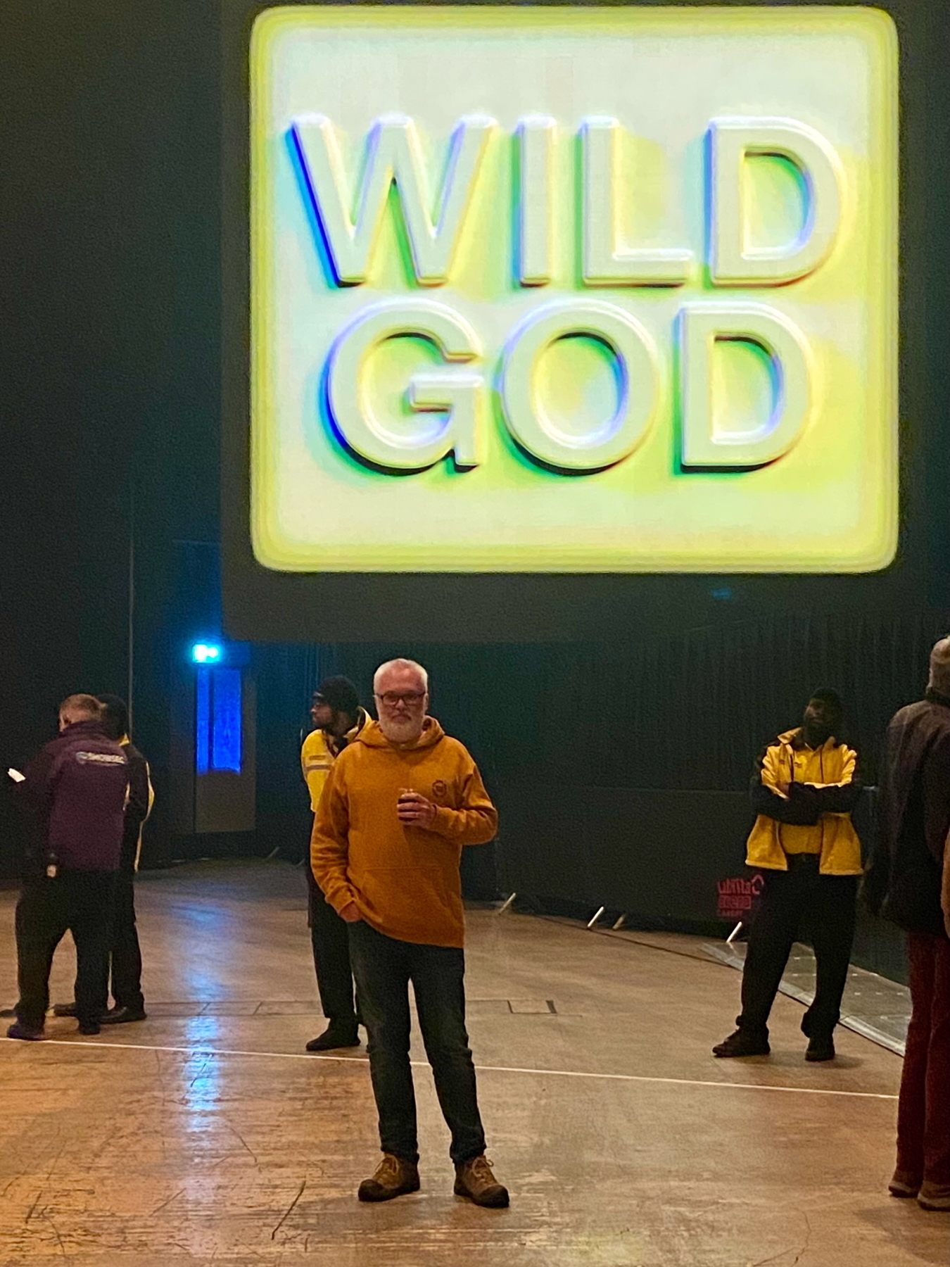 A man wearing a yellow hoodie stands with a drink in a large indoor venue. Behind him, there's a large illuminated sign reading WILD GOD. Several security personnel are visible in the background. The setting appears to be an event or concert.