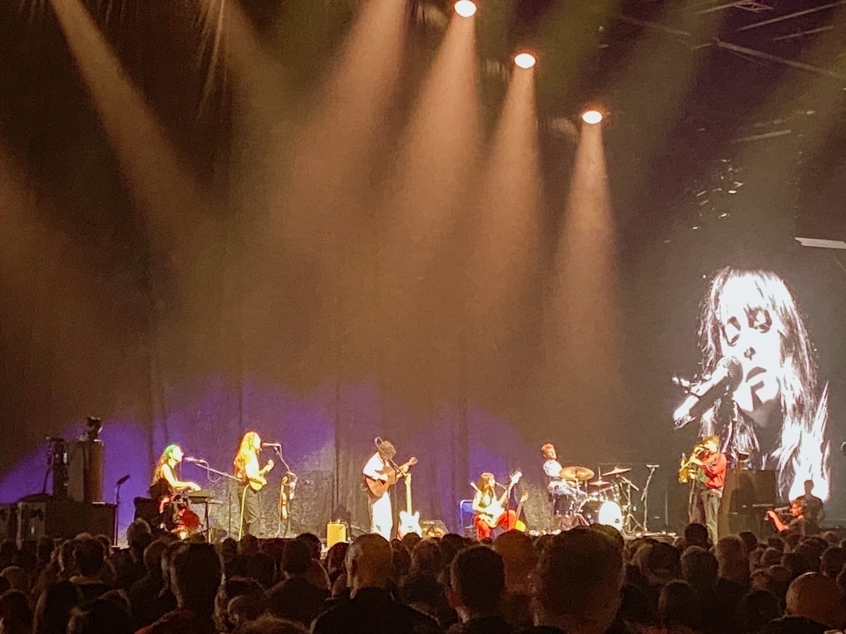 A band performing on stage with multiple musicians playing instruments. There is dramatic stage lighting and a large screen displaying a close-up of a singer. The audience is visible in the foreground.