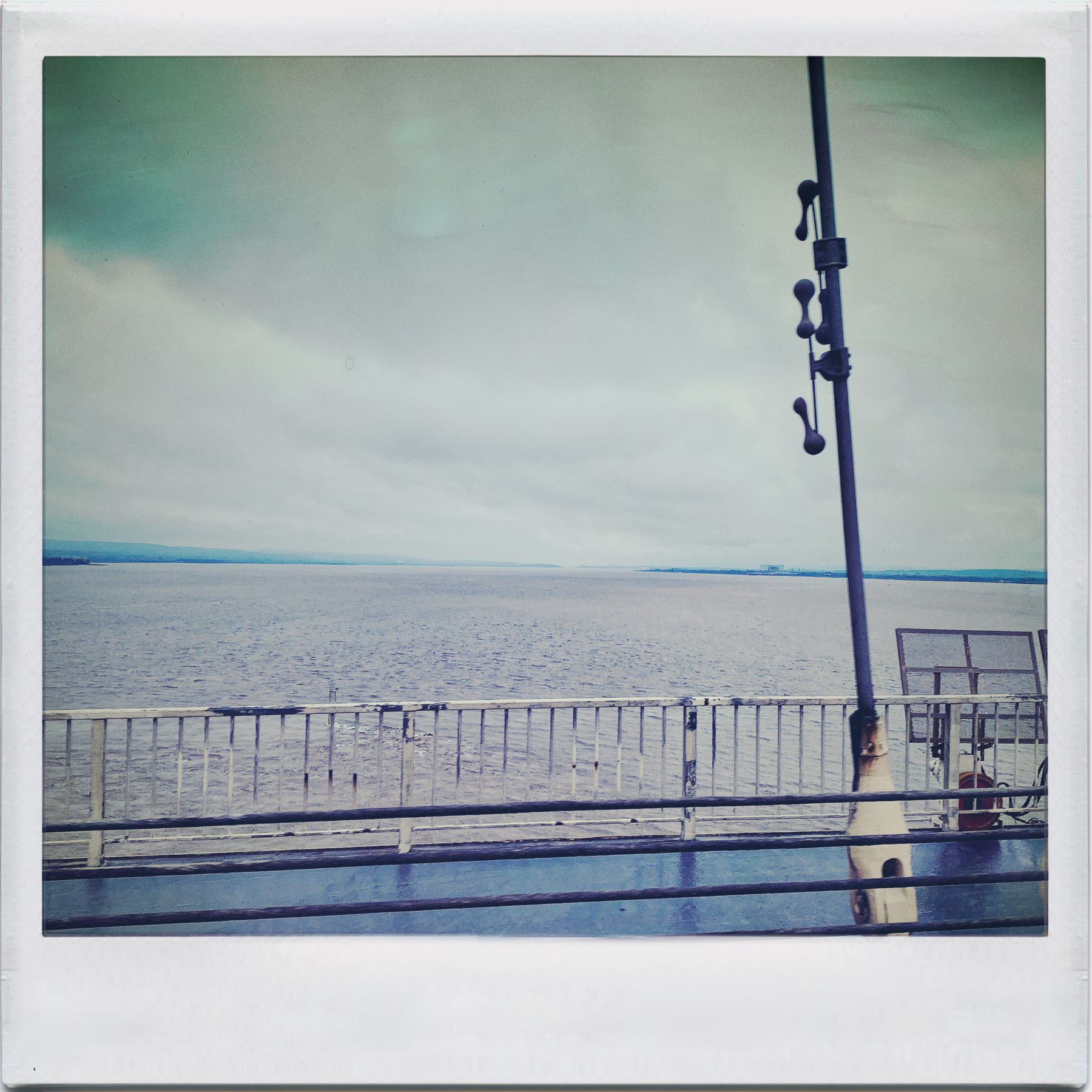 A vintage-style photograph of a sea view from a pier with a metal railing and a lamp post, under an overcast sky.