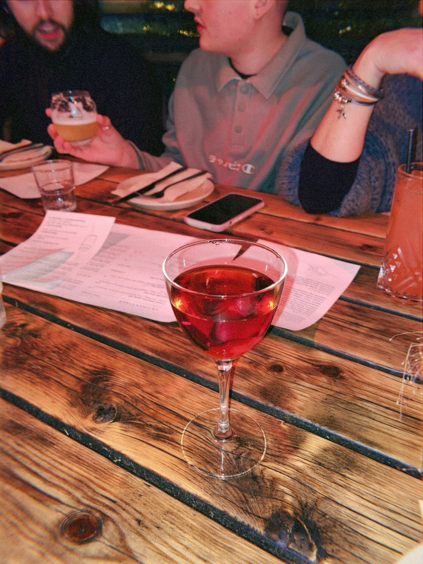 A cozy setting with people enjoying drinks around a wooden table, which features a glass of red beverage in the foreground, and various items such as more drinks, a smartphone, and a menu.