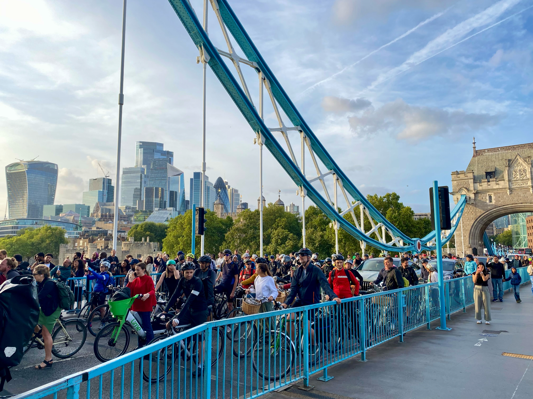 A bustling scene on Tower Bridge in London, showing a large group of cyclists and pedestrians during what appears to be a busy day. The iconic modern London skyline, including The Gherkin and The Walkie Talkie building, is visible in the background