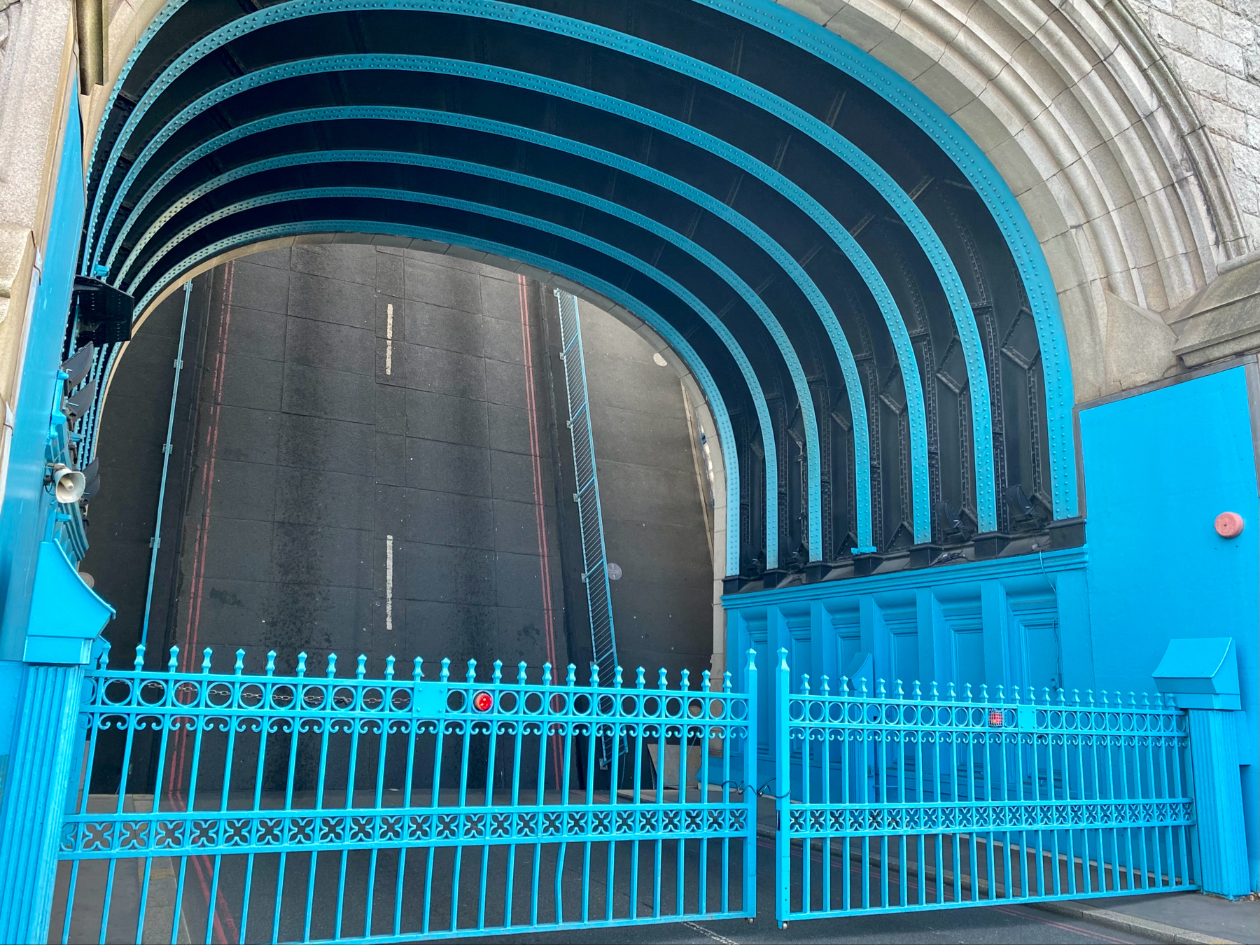 A close-up view of the iconic blue ironwork and gate of London's Tower Bridge, with the raised road visible in the background. The structure displays the distinct blue and black arches typical of the bridge's design.