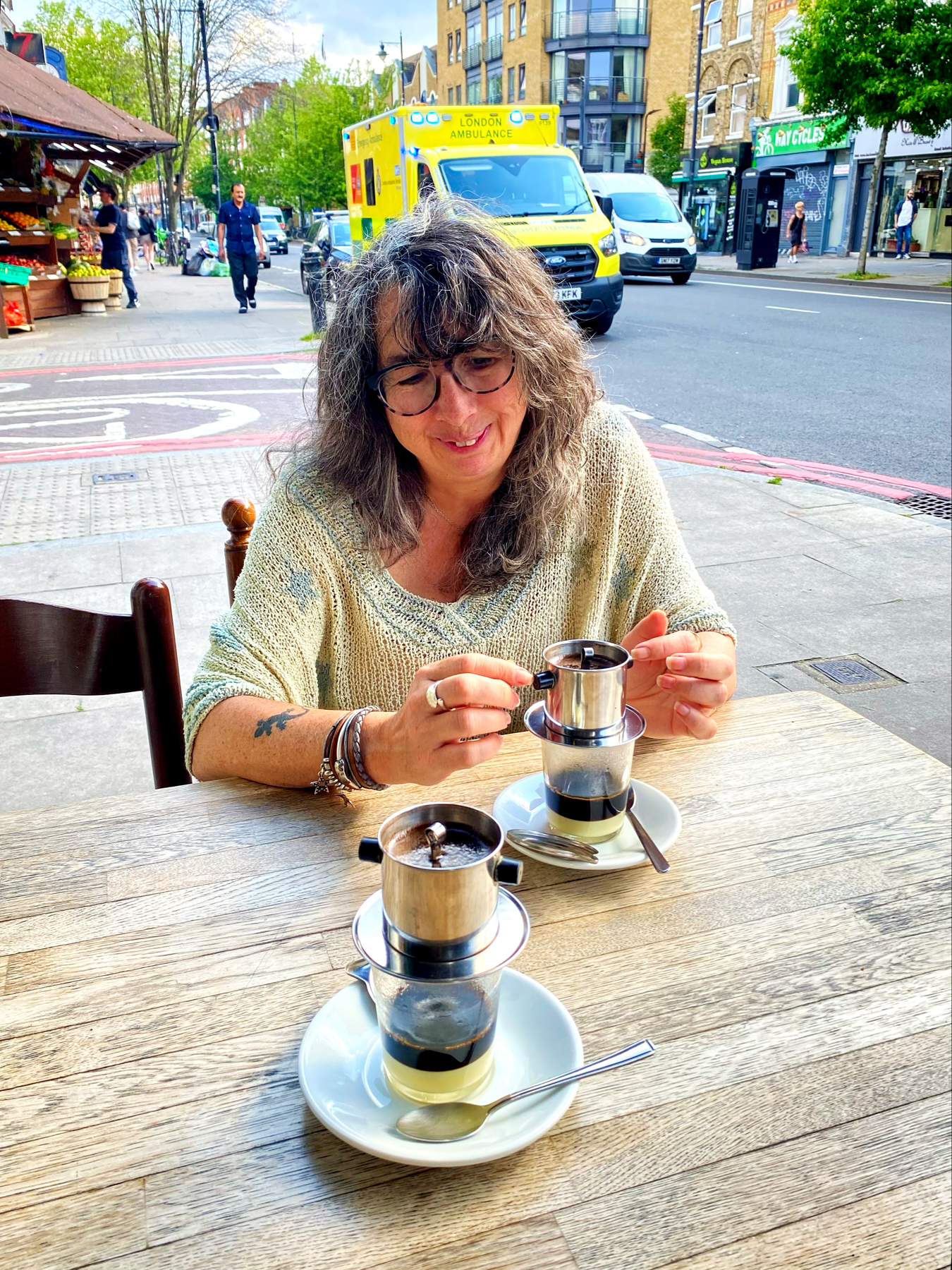 A woman with glasses is sitting at an outdoor café table, smiling and preparing Vietnamese coffee using a traditional drip filter. Two glasses of coffee with condensed milk are on the table. In the background, a London ambulance and a few buildings are visible.