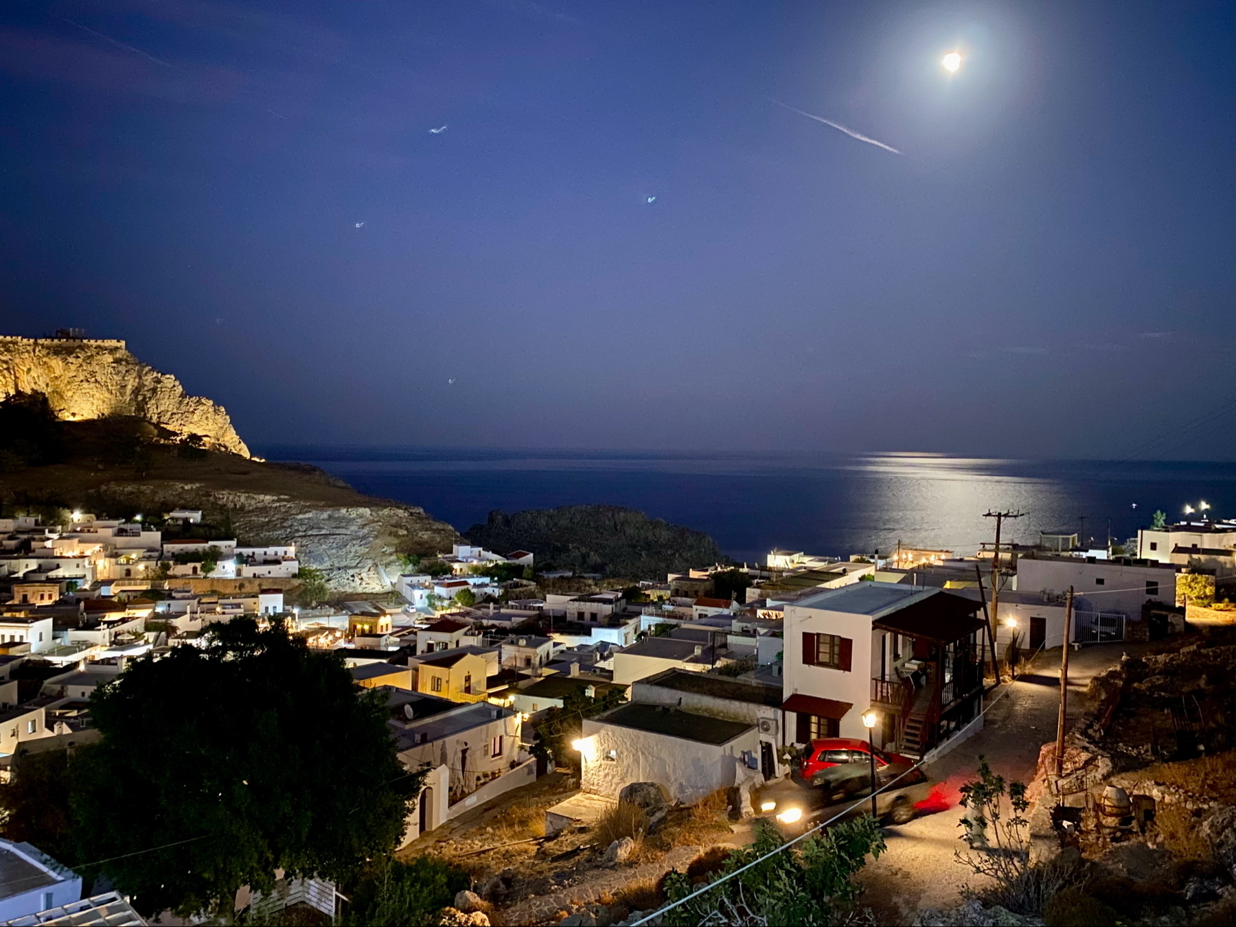 A moonlit coastal village scene at night, featuring illuminated white buildings and a rocky hillside. The sea shimmers under the moonlight, and a car's headlights create light trails along a narrow road.