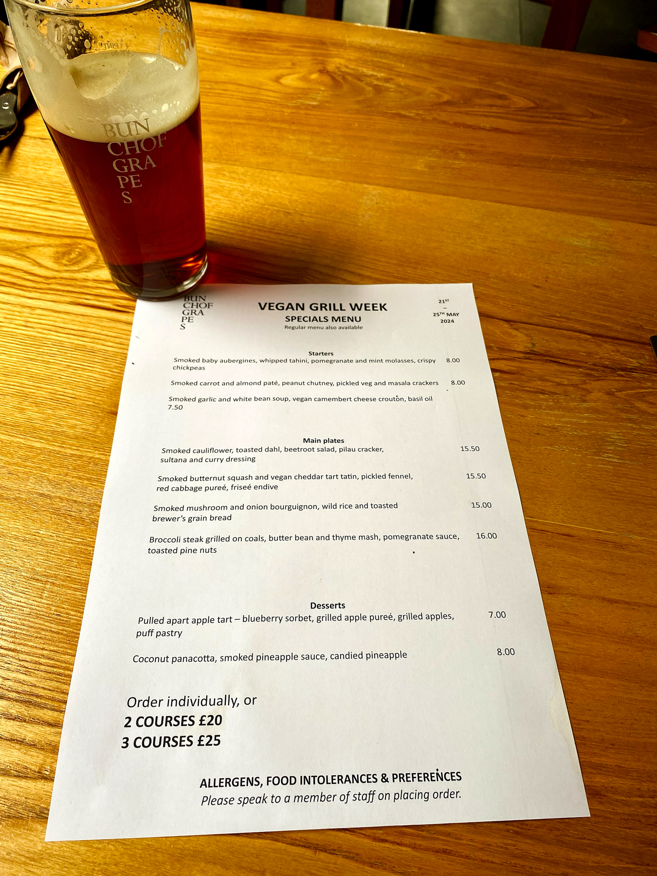 A pint of amber beer sits on a wooden table next to a printed menu for Vegan Grill Week, running from May 21st to May 25th, 2024. The menu features a selection of smoked and grilled vegetable dishes.