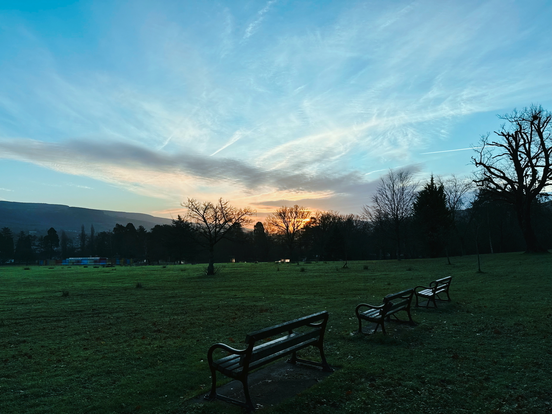 A serene park scene features empty benches on a grassy field with trees silhouetted against a sunrise sky.