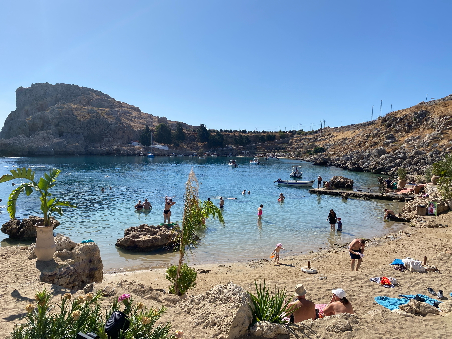 A sunny beach scene with people swimming and relaxing on the sand. There's a rocky shoreline and clear blue water. Boats are anchored in the bay, and potted plants are on the beach. The backdrop features rocky hills under a clear sky.