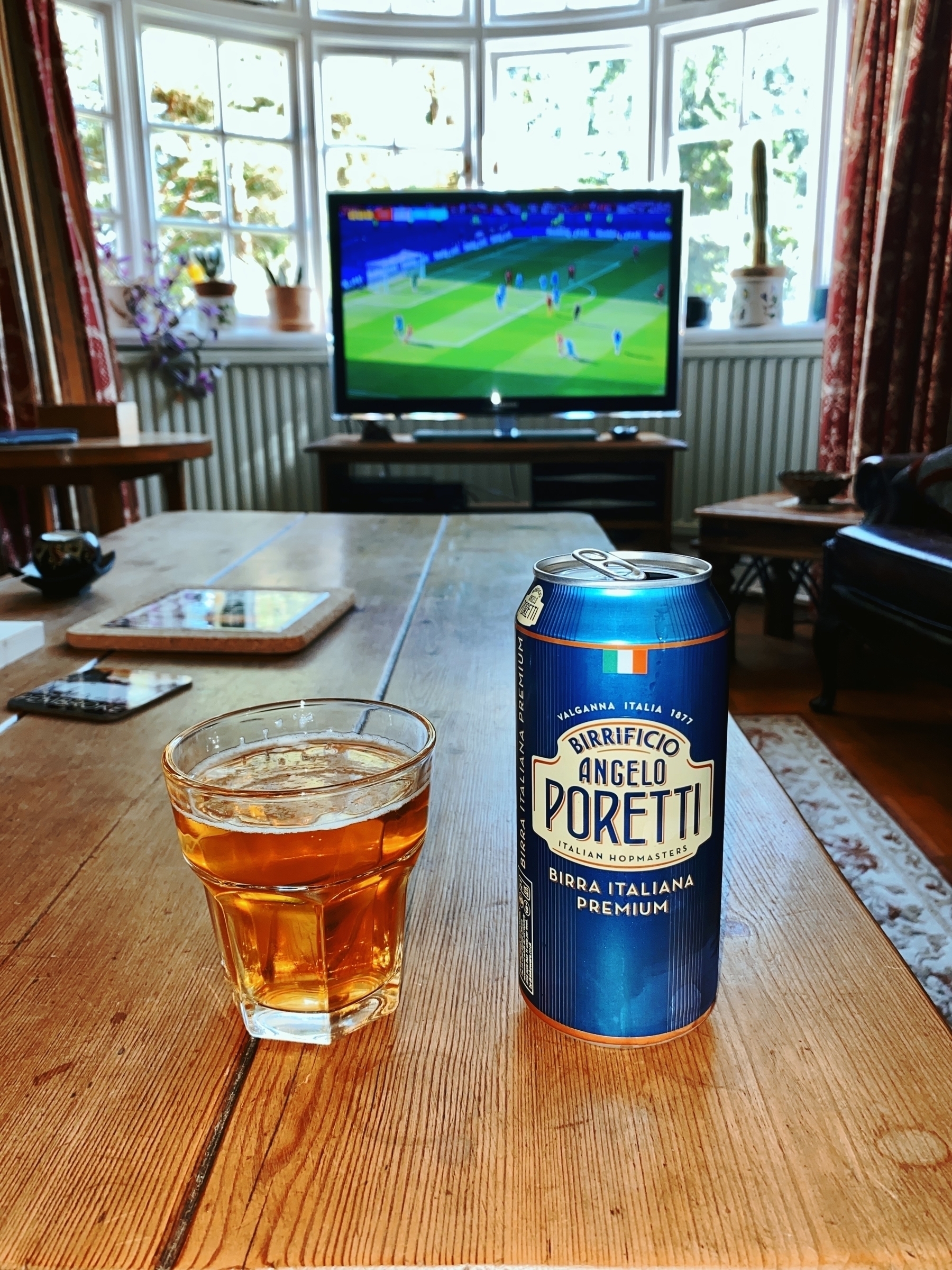 A glass of beer and a can of Birrificio Angelo Poretti sit on a wooden table in a living room with a soccer game on TV in the background.