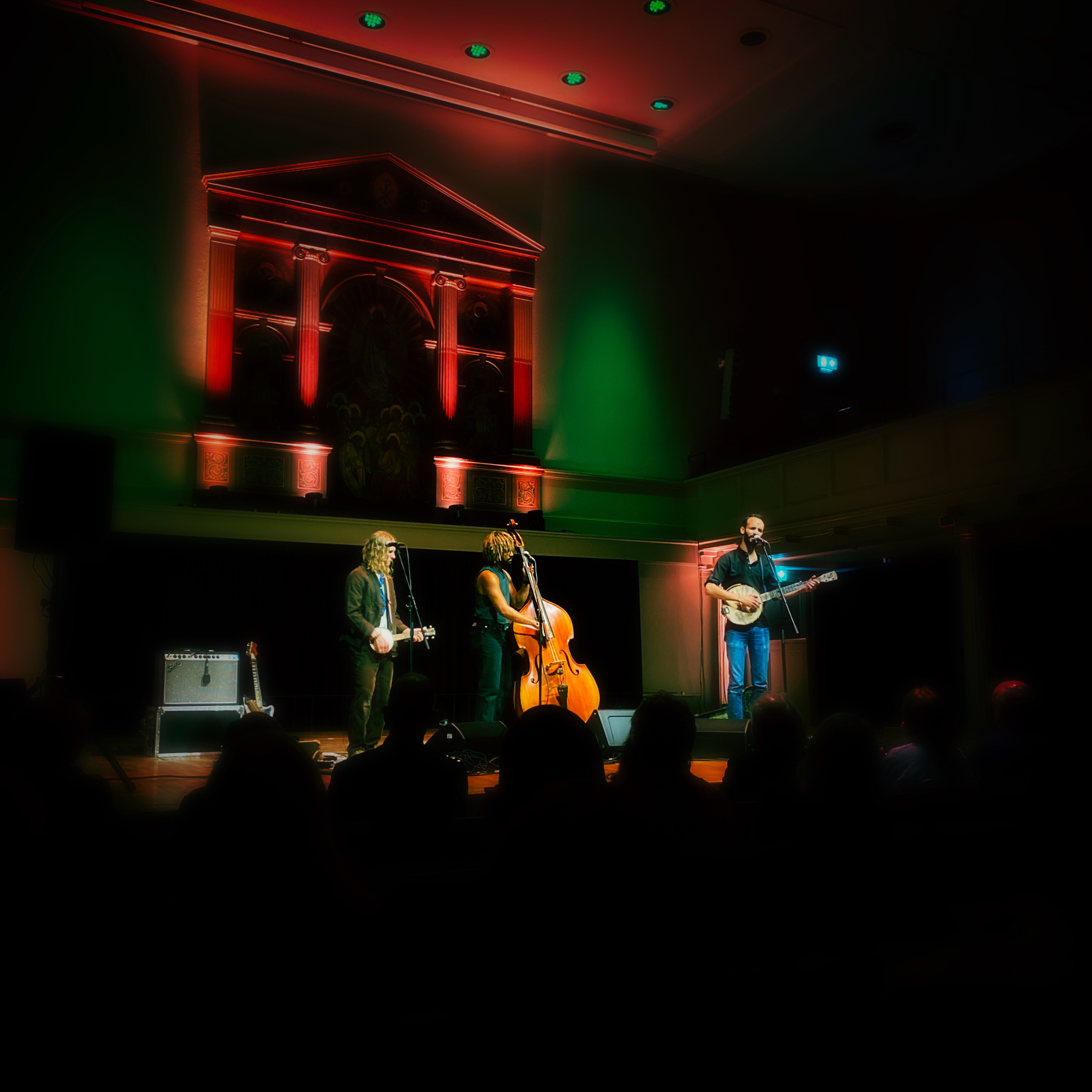 A musical trio performing on stage in a dimly-lit venue with colorful lighting. The three musicians are playing different instruments, including a banjo and a double bass, with a backdrop featuring architectural elements lit in red and green.