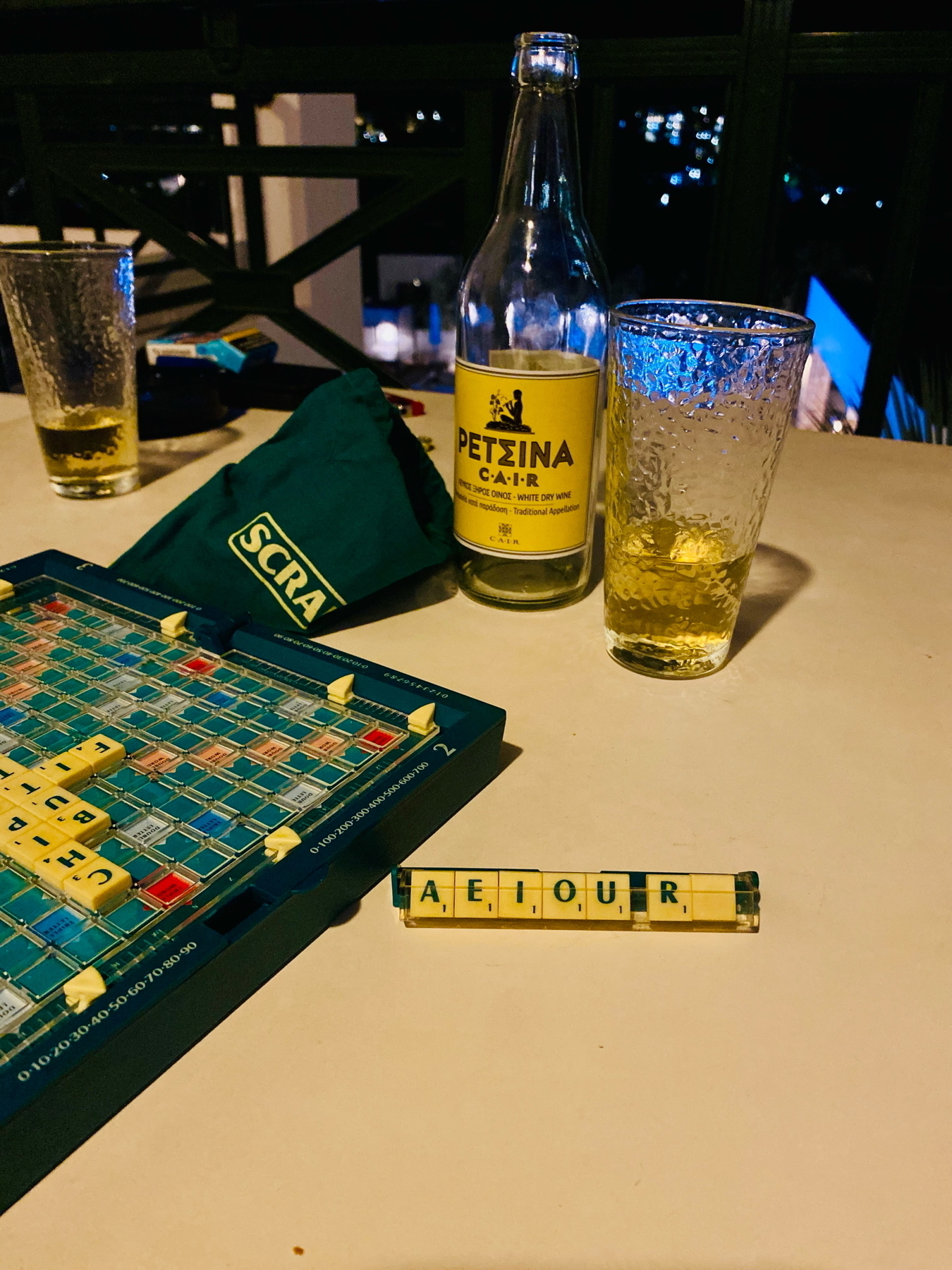 A Scrabble board game setup on a table with a rack of tiles showing the letters A, E, I, O, U, R and a blank tile. A bottle labeled CAIR Retsina and a partially filled glass are nearby.