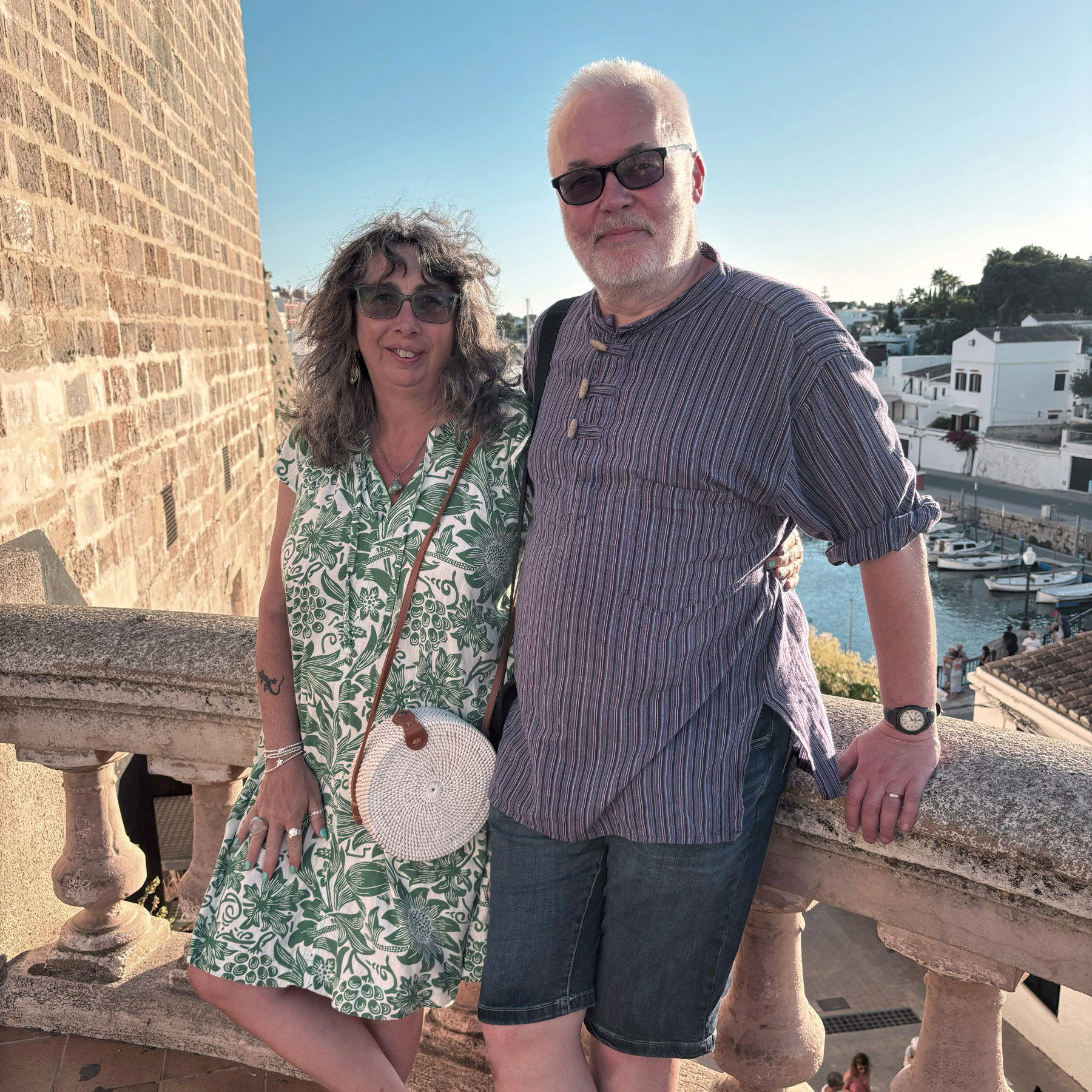A couple standing together, posing for a photo on a stone terrace with a scenic backdrop of a coastal town. The man is wearing sunglasses, a striped shirt, and shorts, while the woman is in a green patterned dress with a white woven handbag.