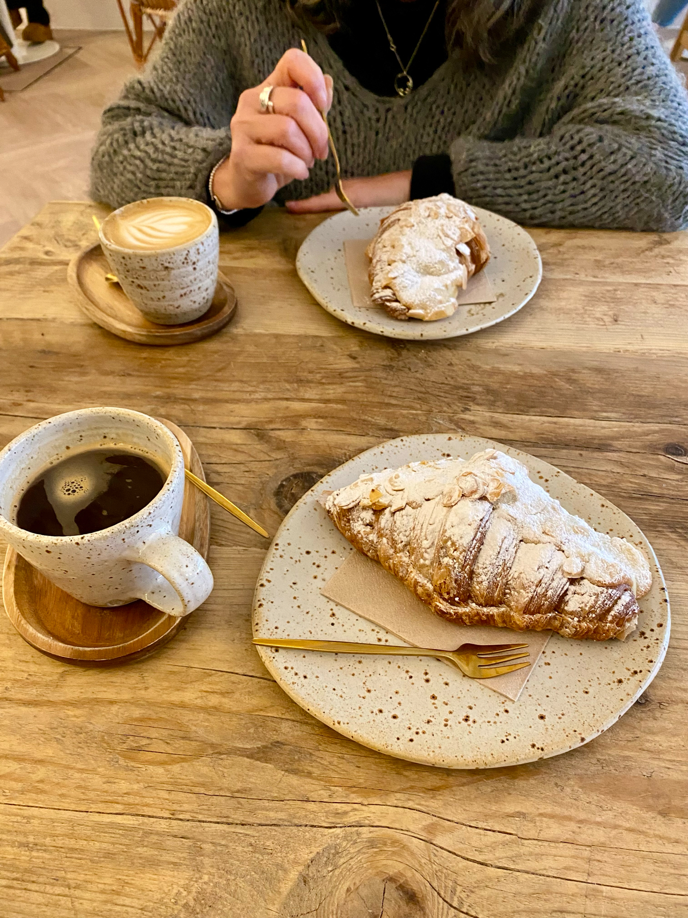 A person seated at a wooden table enjoying a coffee and pastries, including a frothy cappuccino and a dusted almond croissant.